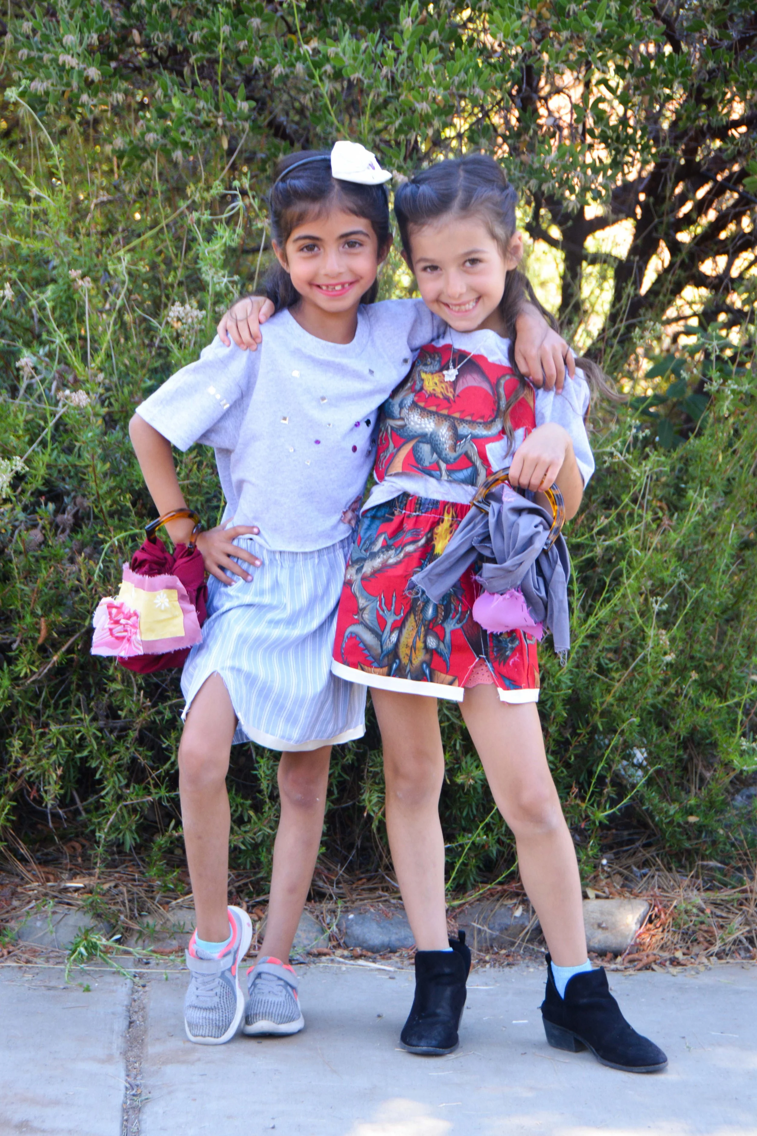 Two young girls standing outdoors on sidewalk, smiling, with their arms around each other, surrounded by greenery.