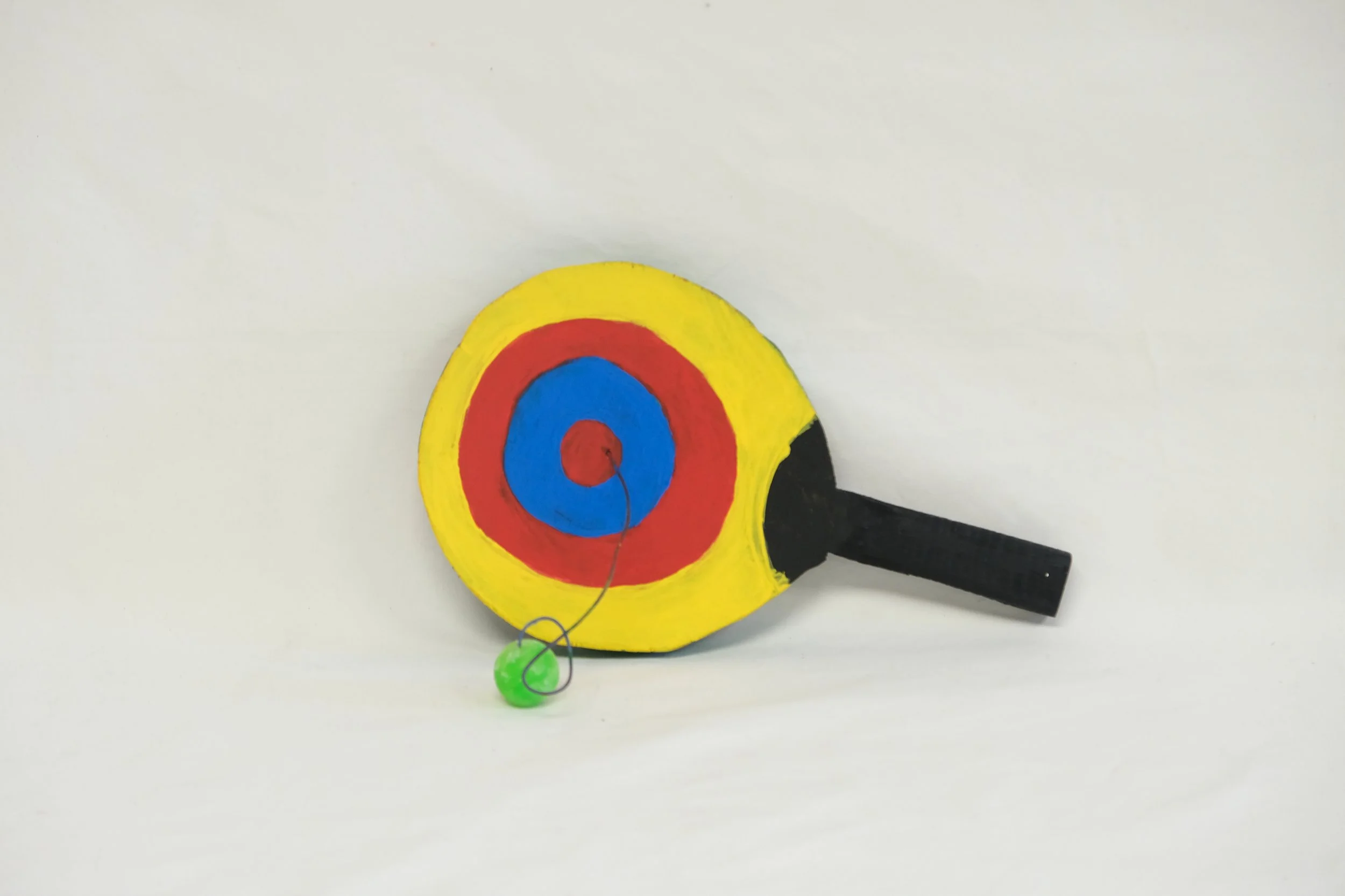 Handmade toy dartboard with a black handle, painted yellow, red, and blue concentric circles, and a green dart attached with a string.