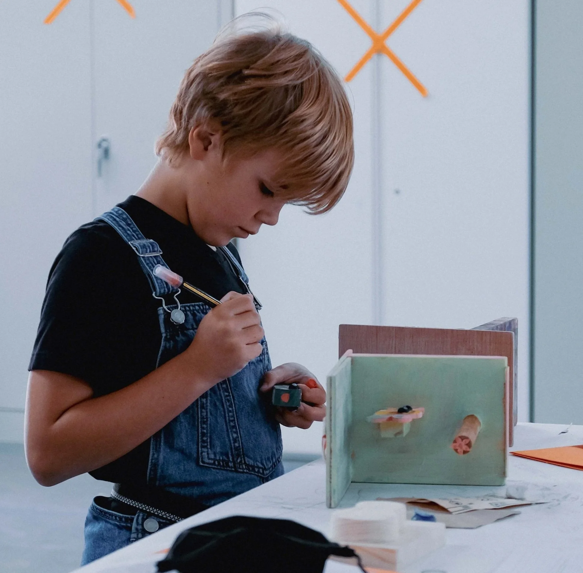A young boy with blonde hair, wearing a black t-shirt and denim overalls, is standing at a table working on a craft project with paper and markers inside a room with storage cabinets and boxes in the background.