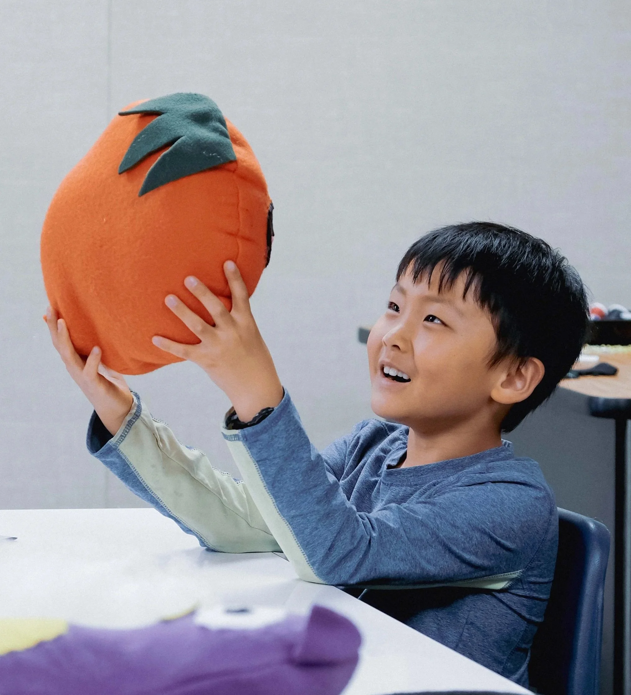 A young boy with dark hair smiling and holding a plush toy shaped like an orange with green leaves on top.