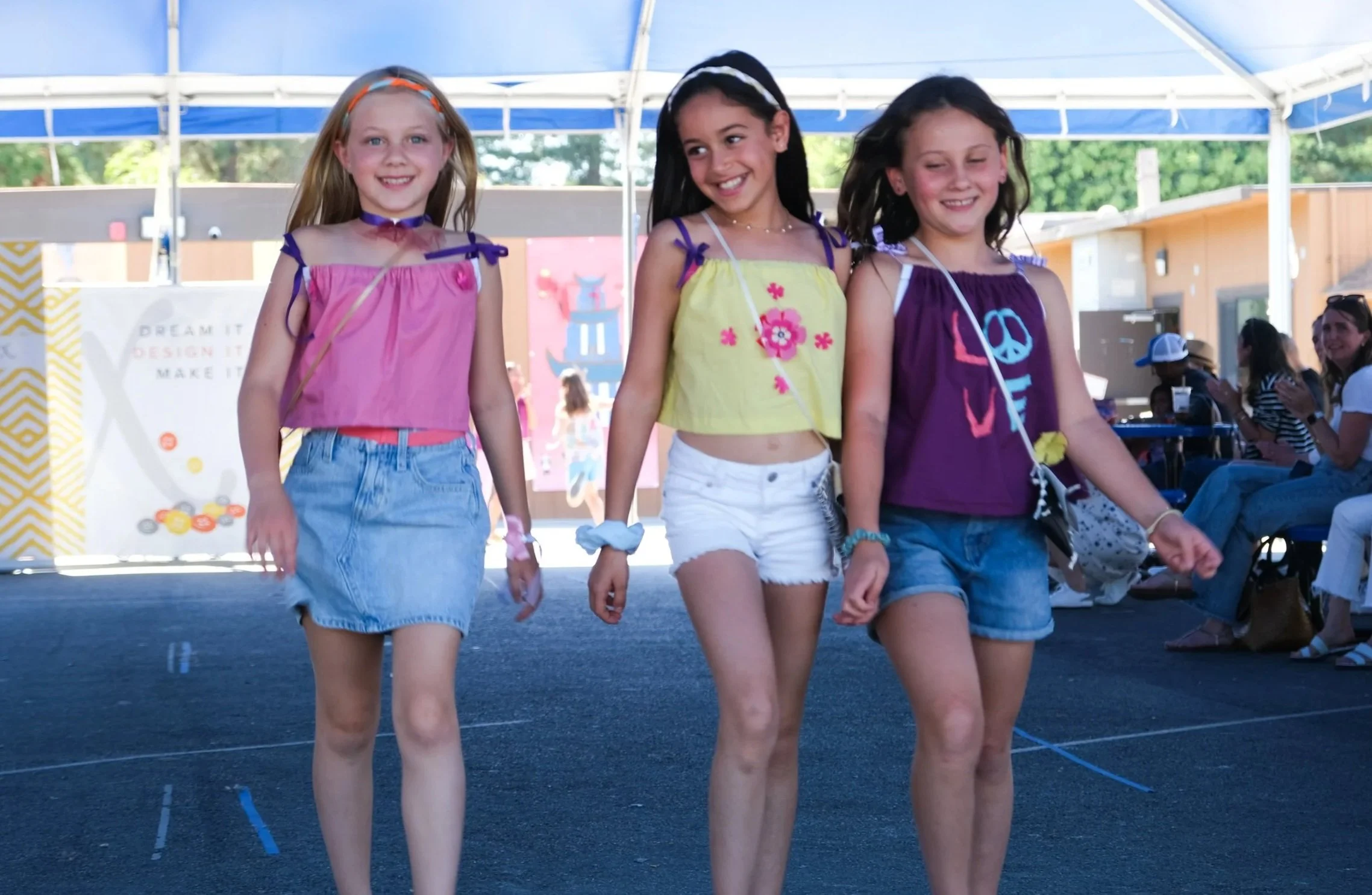 Three young girls walking on a stage with an audience in the background during an outdoor event.