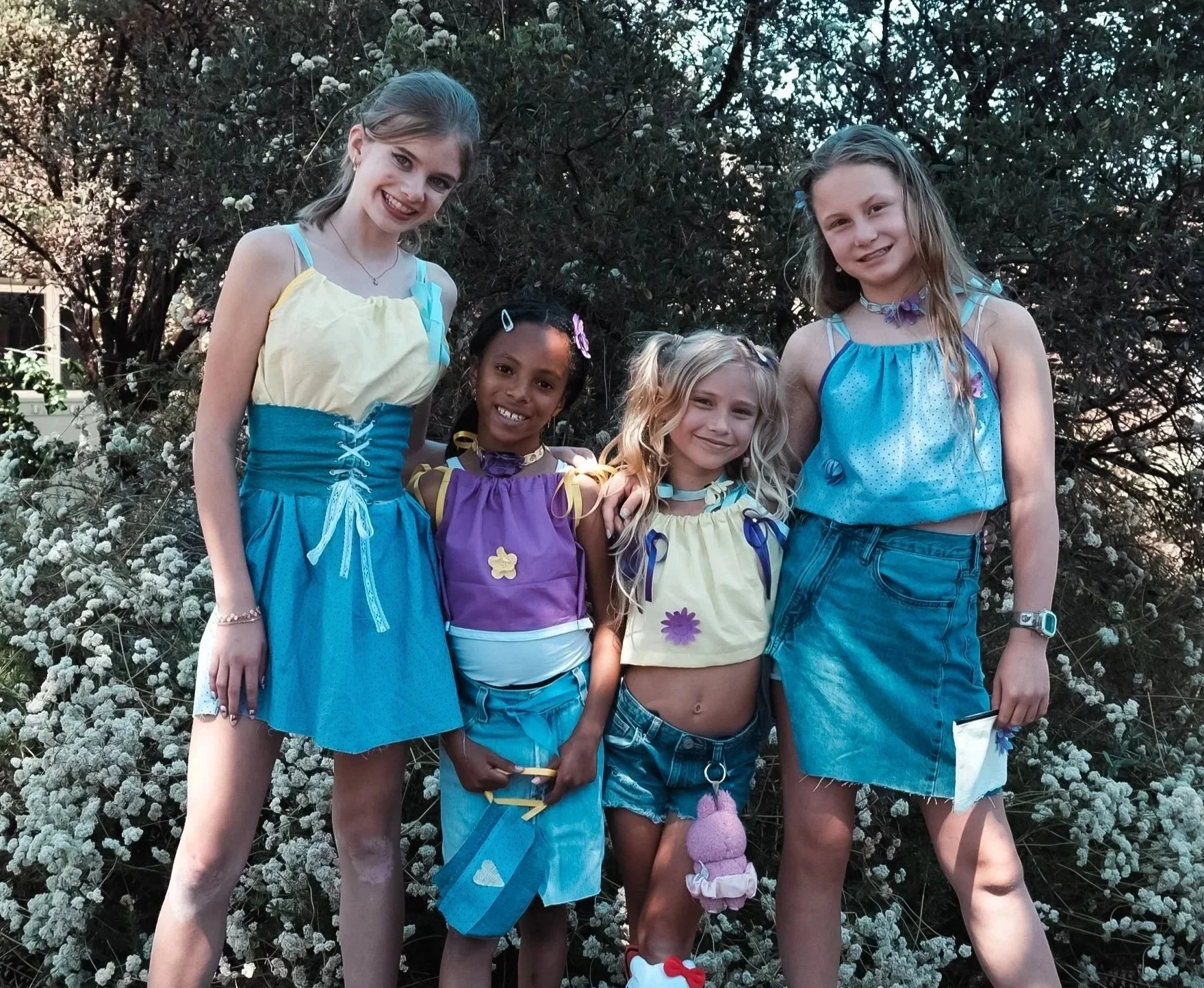 Five young girls standing together outdoors in front of bushes, smiling at the camera. They are wearing casual summer clothing.