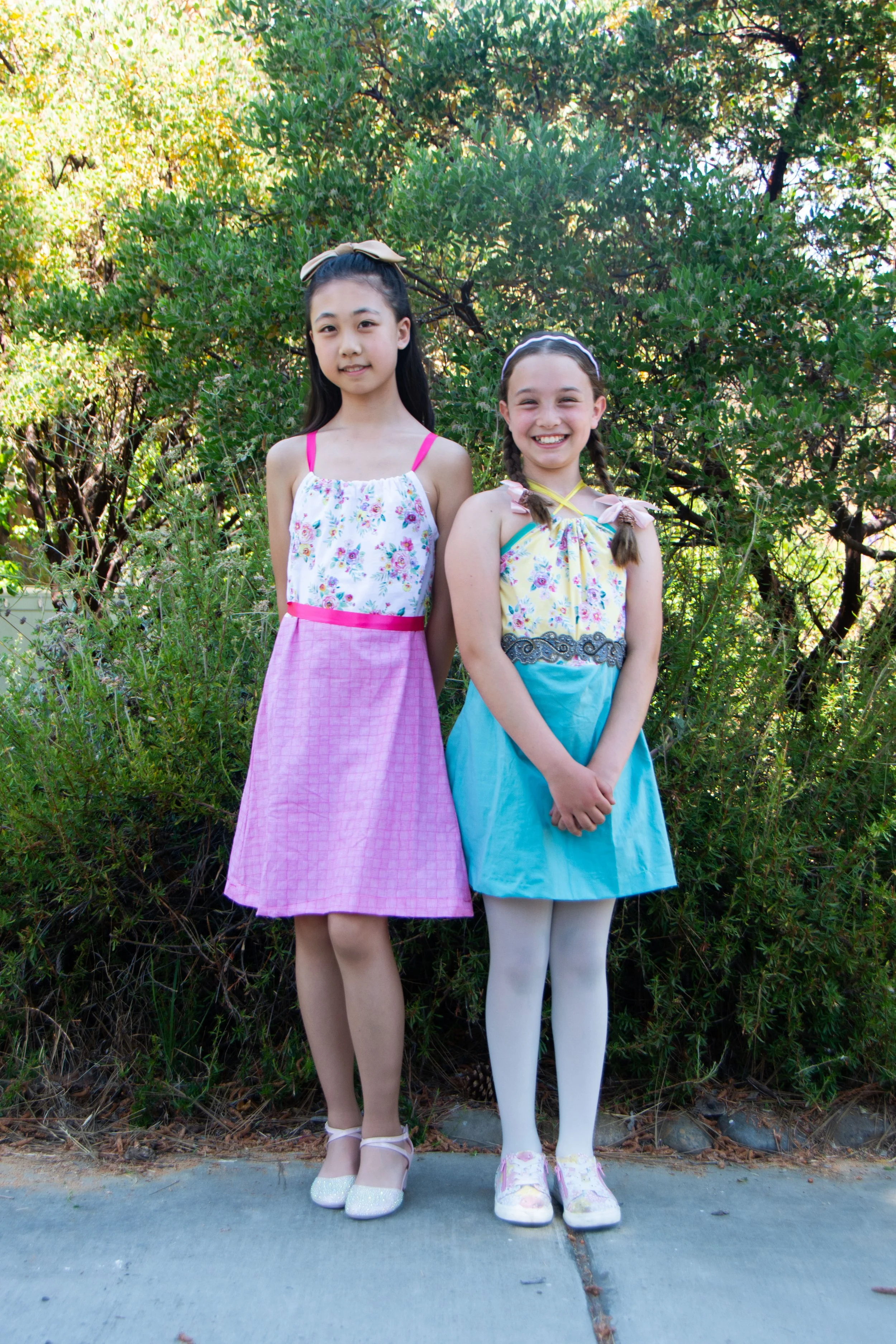 Two young girls standing outdoors in front of green bushes, wearing colorful dresses and smiling.