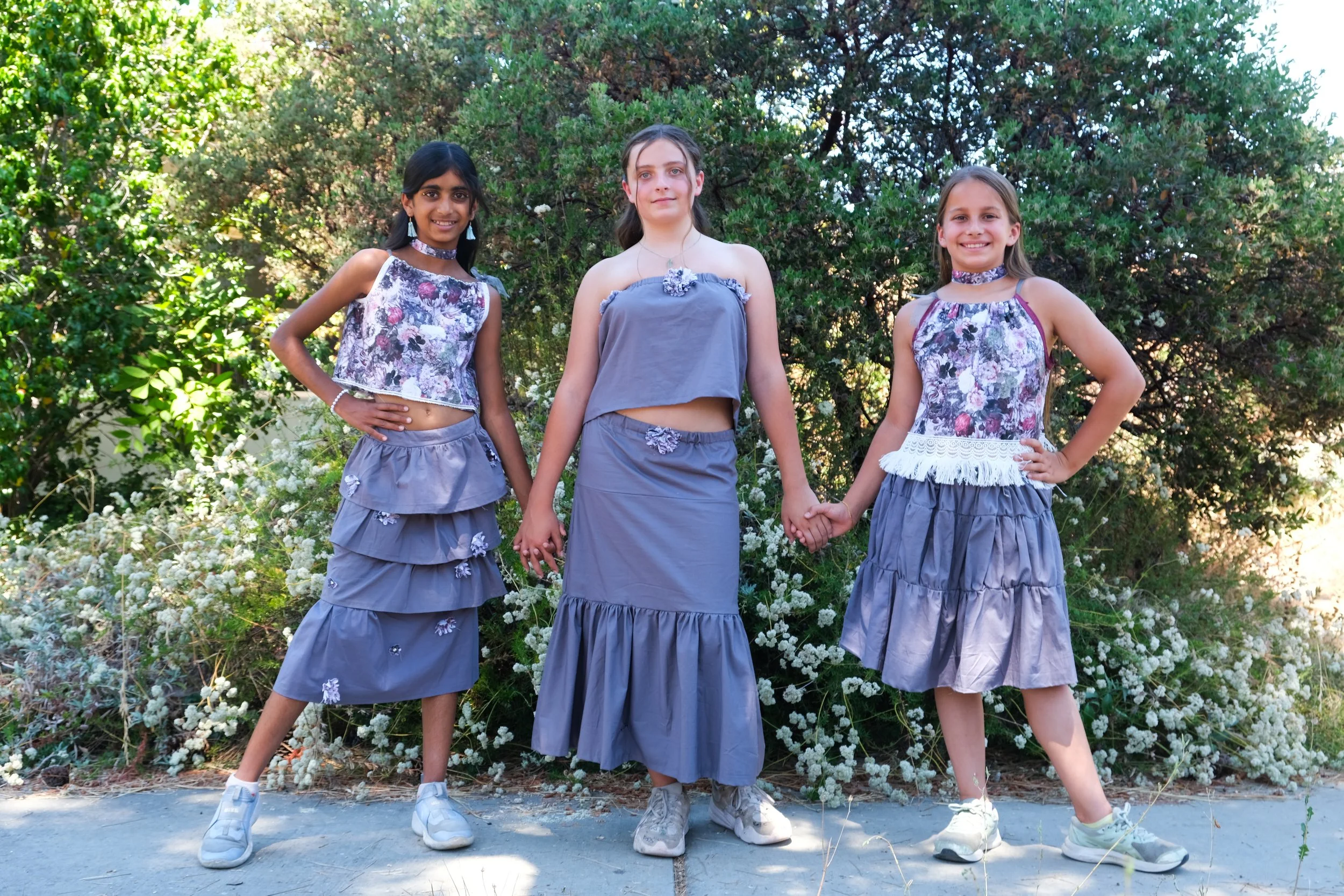Three girls standing outdoors in front of green bushes, holding hands, wearing coordinated gray and purple outfits with floral patterns and ruffled skirts.