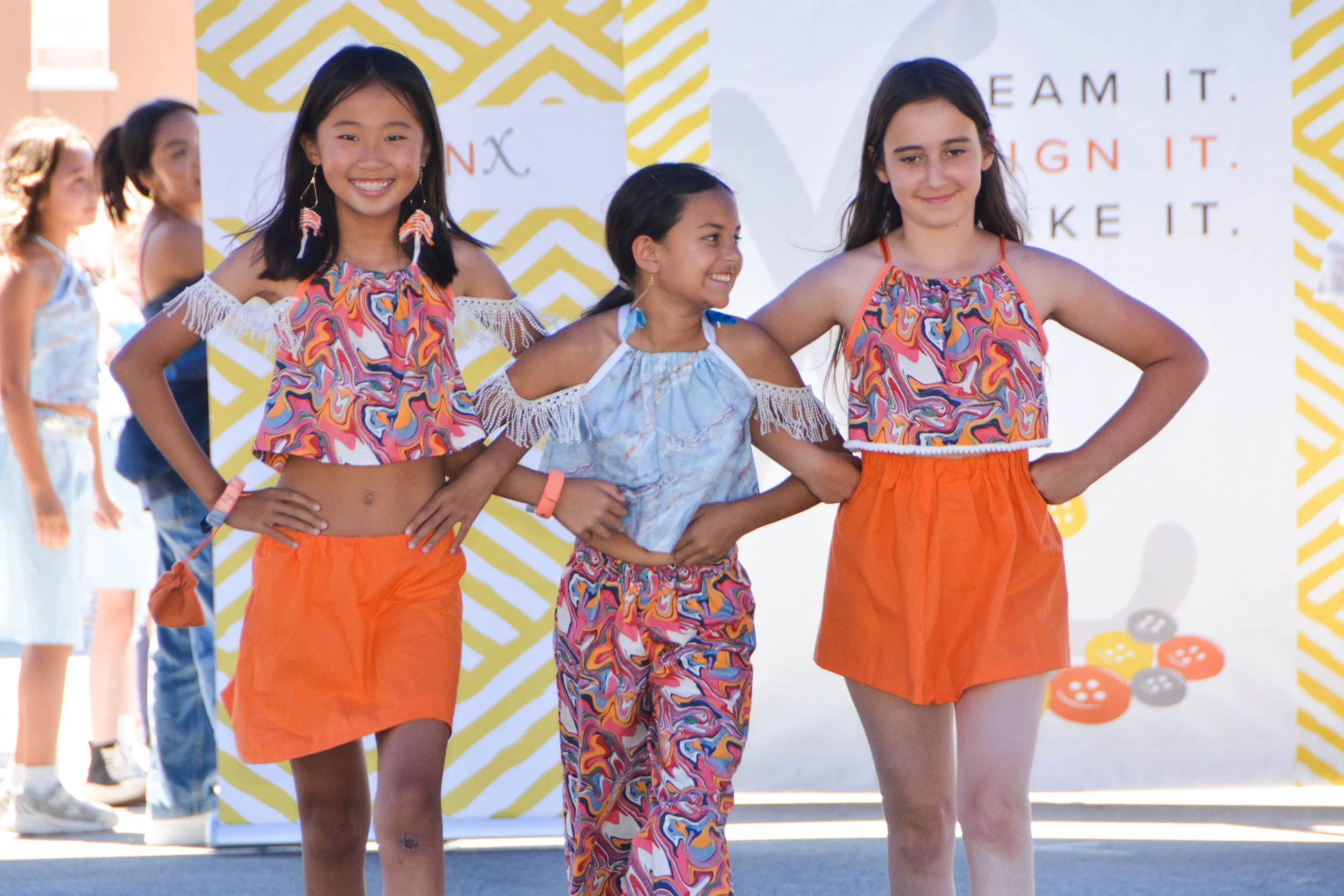 Three young girls on a stage, smiling and holding hands, wearing colorful summer outfits, with other children in the background at an outdoor event.