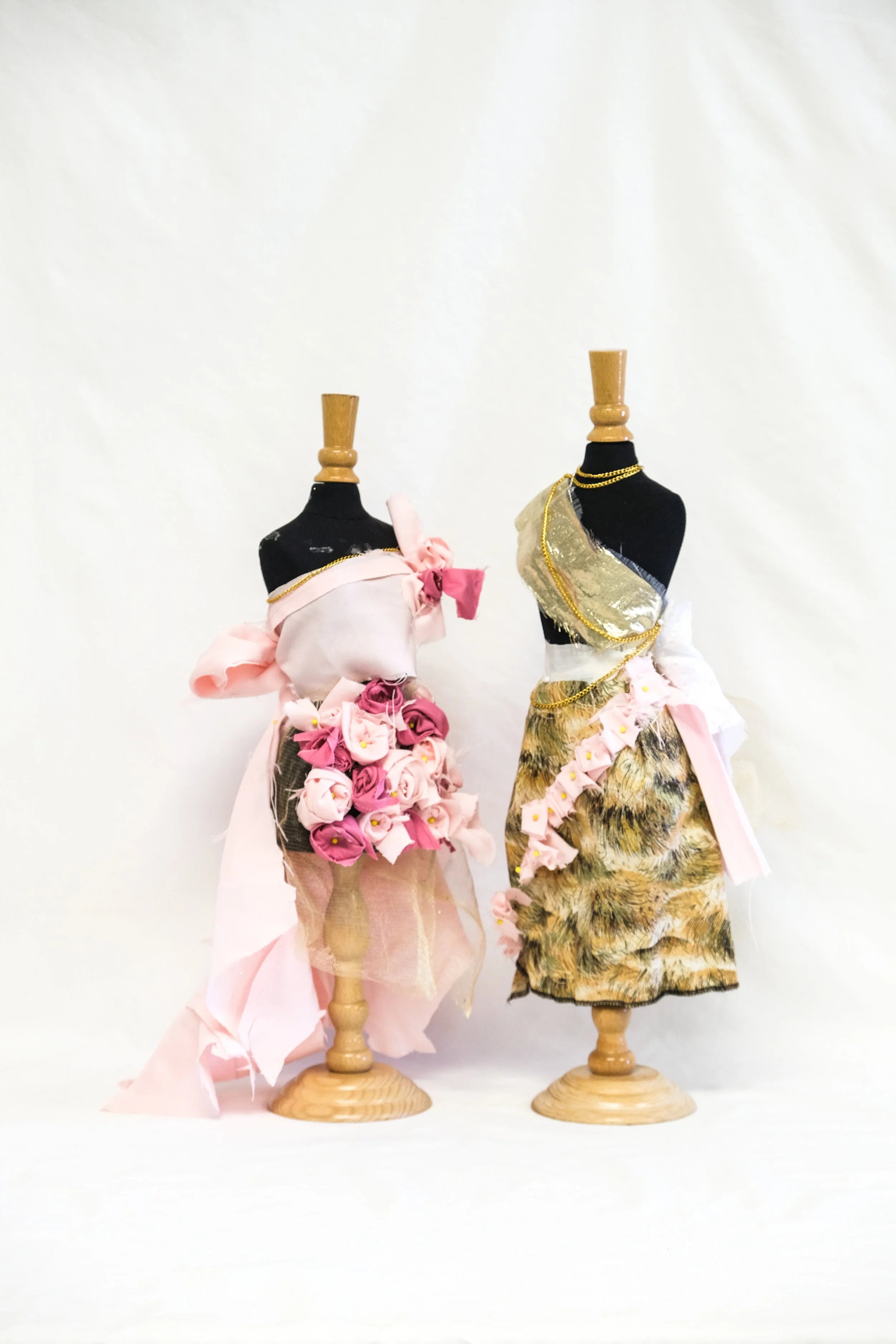 Two dress forms displaying elaborate, artistic fashion designs made from fabric, ribbons, and decorative elements, set against a plain white background.