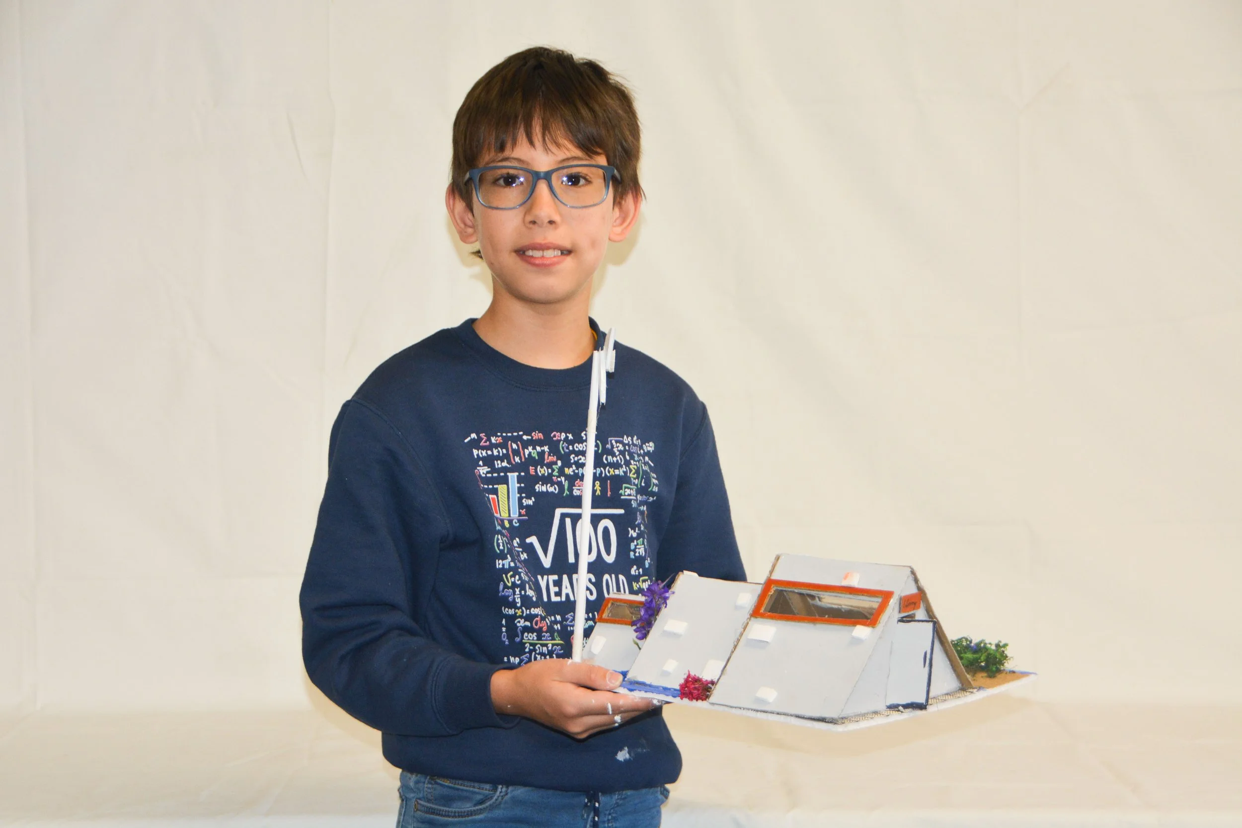 A young boy with glasses and a navy sweatshirt holding a detailed architectural model of a modern house.