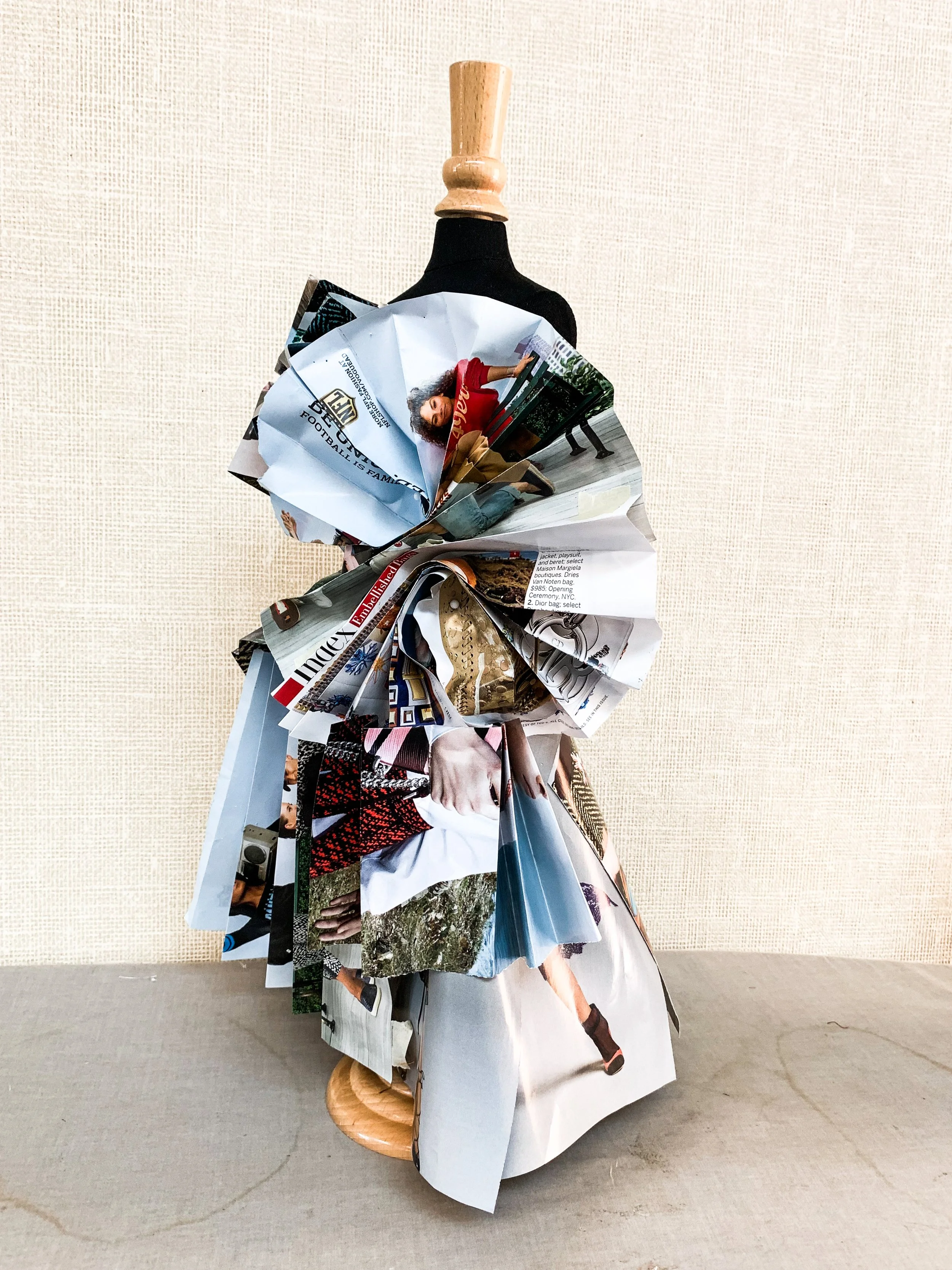 A dressmaker's mannequin draped with a dress made from crumpled and folded magazine pages, displayed on a light-colored surface against a textured beige wall.