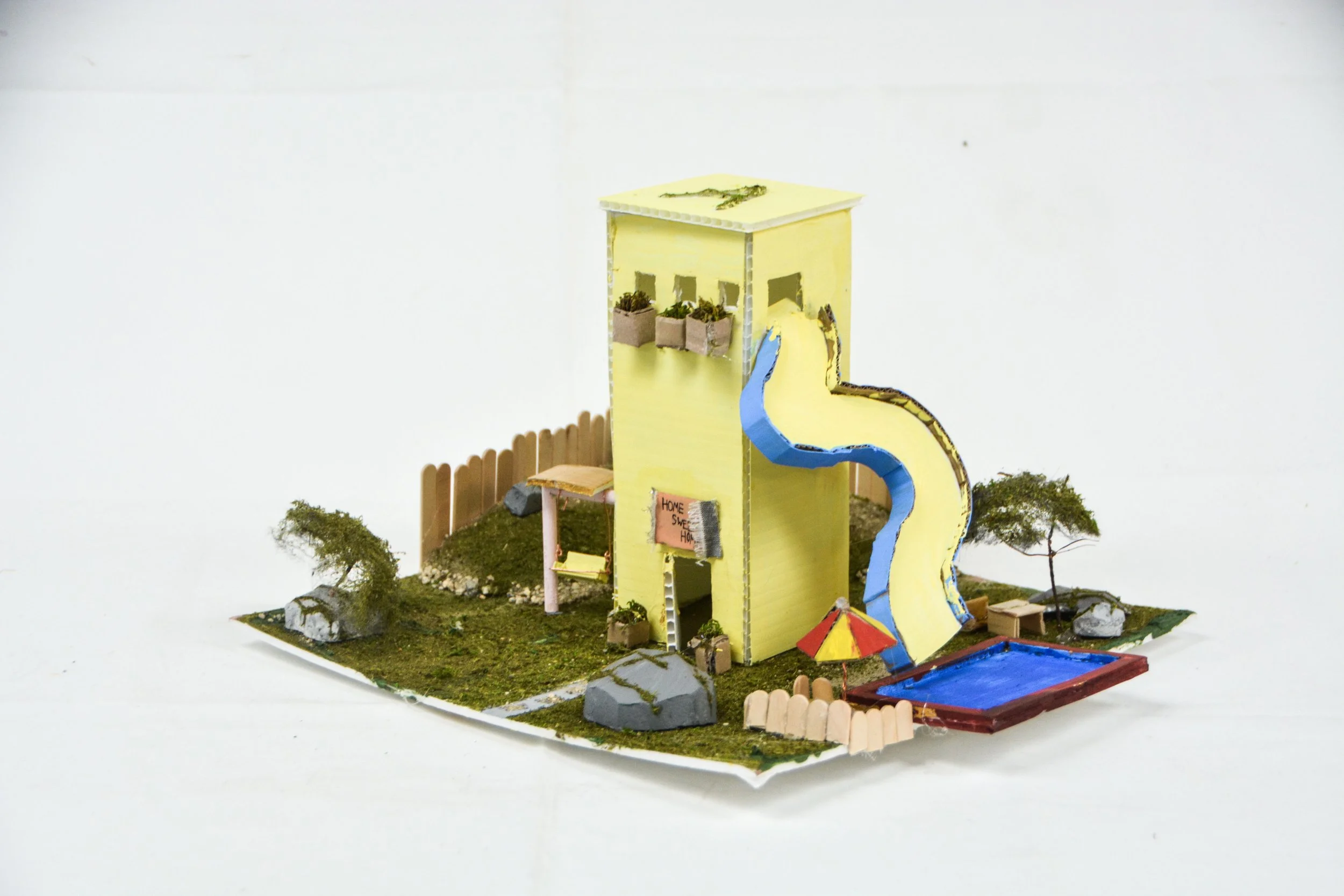 Miniature model of a yellow house with a blue slide, small pool, trees, rocks, and a fence, on a grassy base.