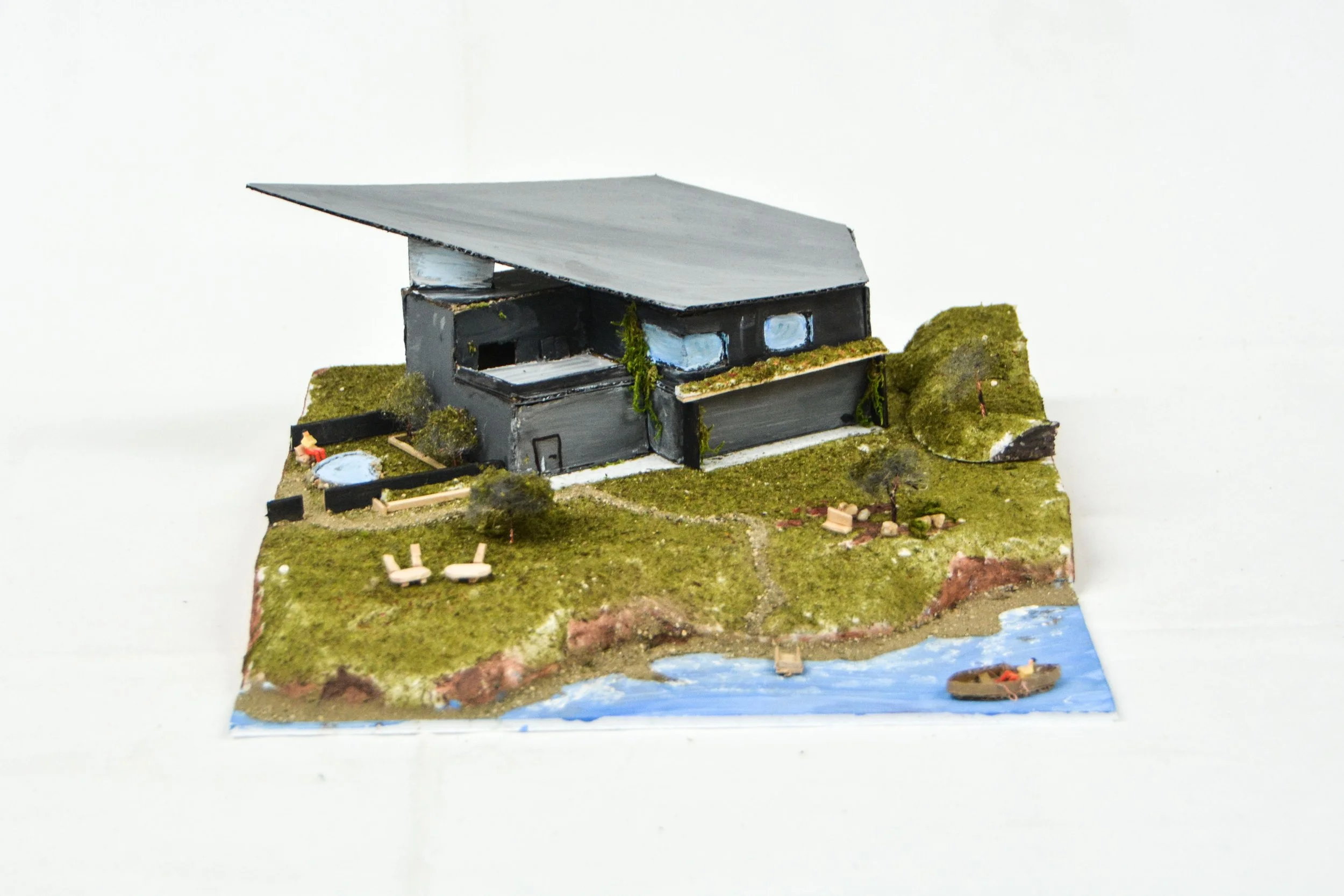 A detailed architectural model of a modern house with a large sloped roof set on a grassy landscape near a water body, featuring trees, pathways, and outdoor furniture.