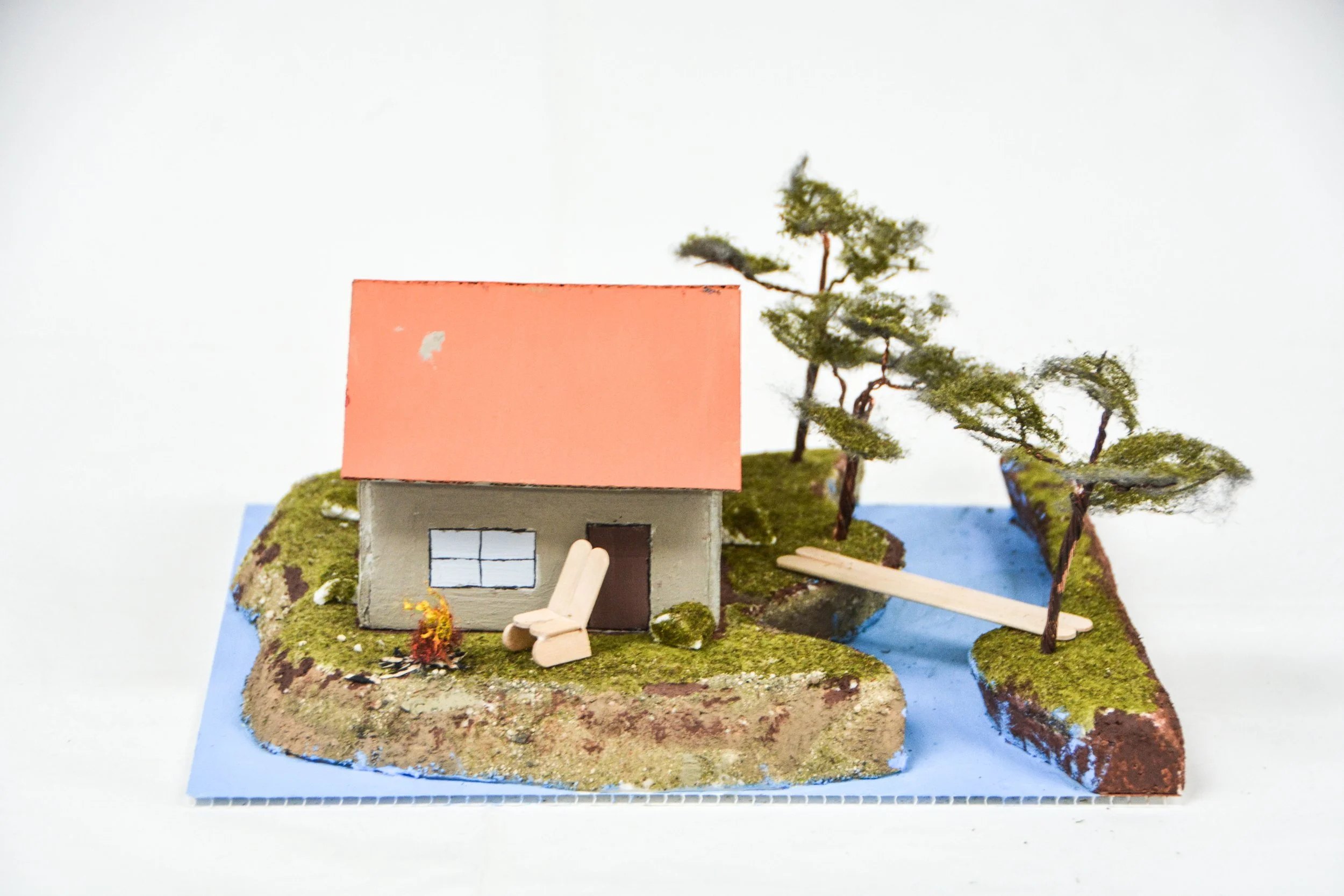 Miniature model of a house with a pink roof, surrounded by trees, on a small terrain with moss and grass, next to a pond with a wooden plank bridge.