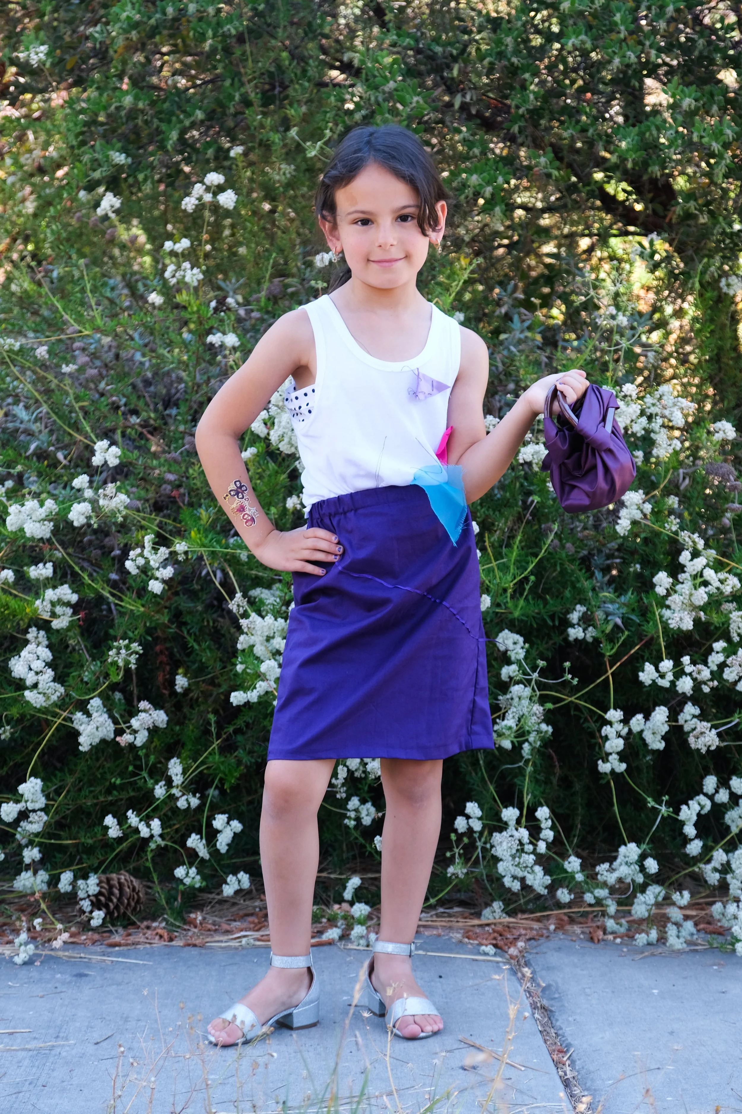 A young girl with dark hair and light skin standing on a sidewalk in front of white flowering bushes, wearing a white sleeveless top, purple skirt, and silver sandals, holding purple clothing in her right hand, with a playful tattoo or temporary tatt