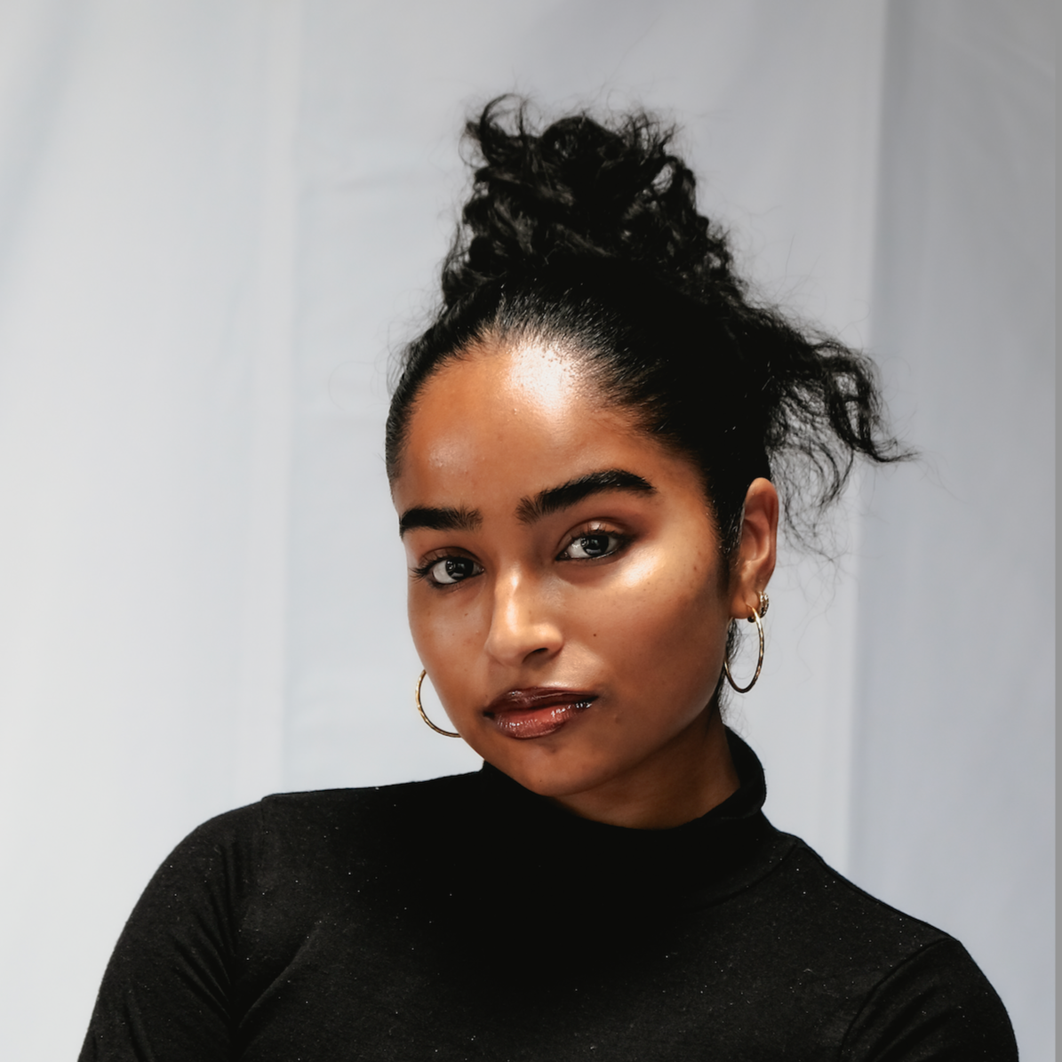 A young woman with dark curly hair styled in a high, messy bun, wearing a black turtleneck and hoop earrings, looking confidently at the camera against a plain background.