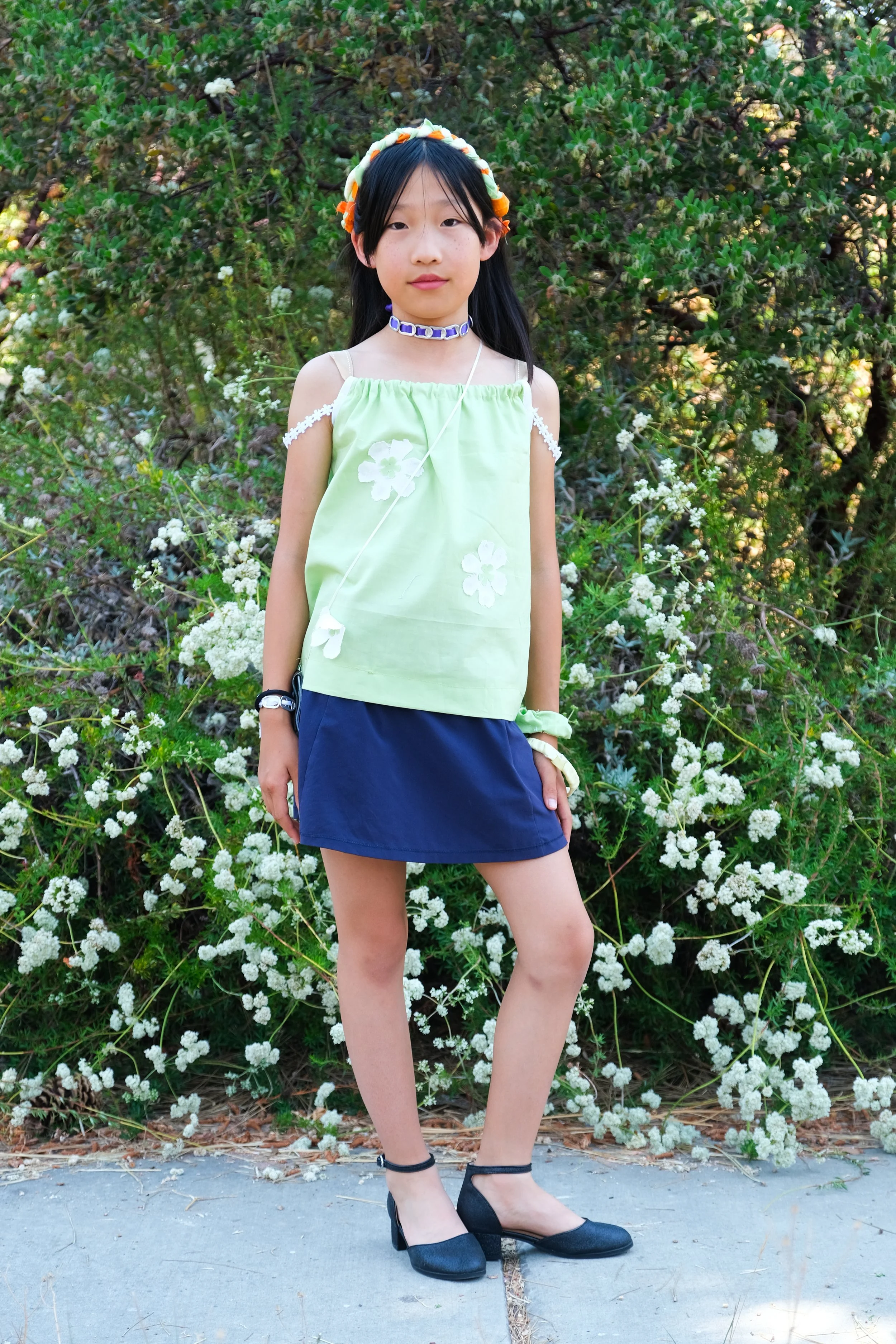 A young girl stands outdoors in front of lush green foliage and white flowers, wearing a light green top with white floral accents, a dark blue skirt, black high heels, and accessories including a headband, necklace, bracelets, and a wristwatch.
