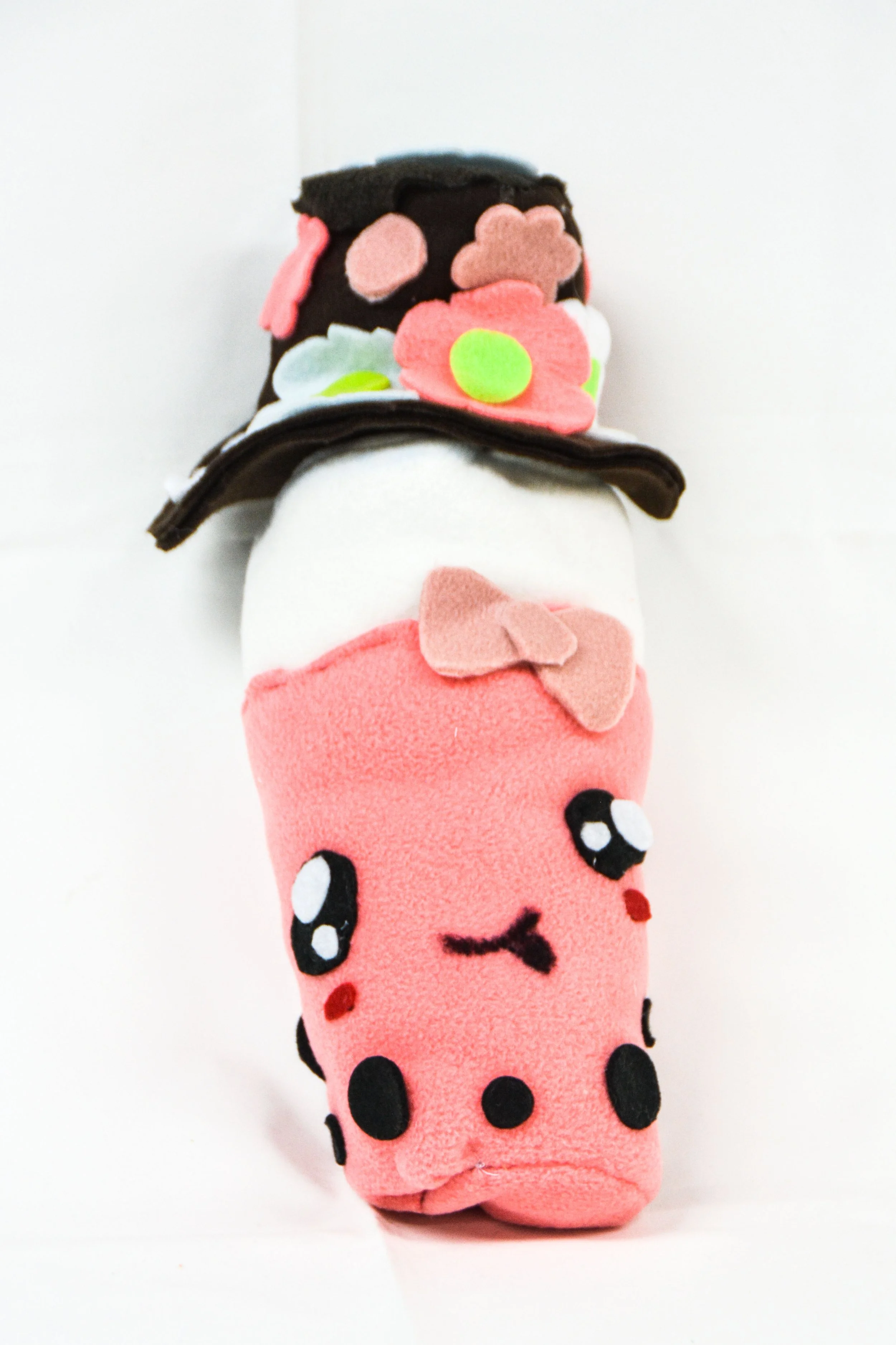 Cute plush toy in the shape of a cupcake wearing a black top hat decorated with colorful felt flowers, with a pink body featuring a cute face and small arms.