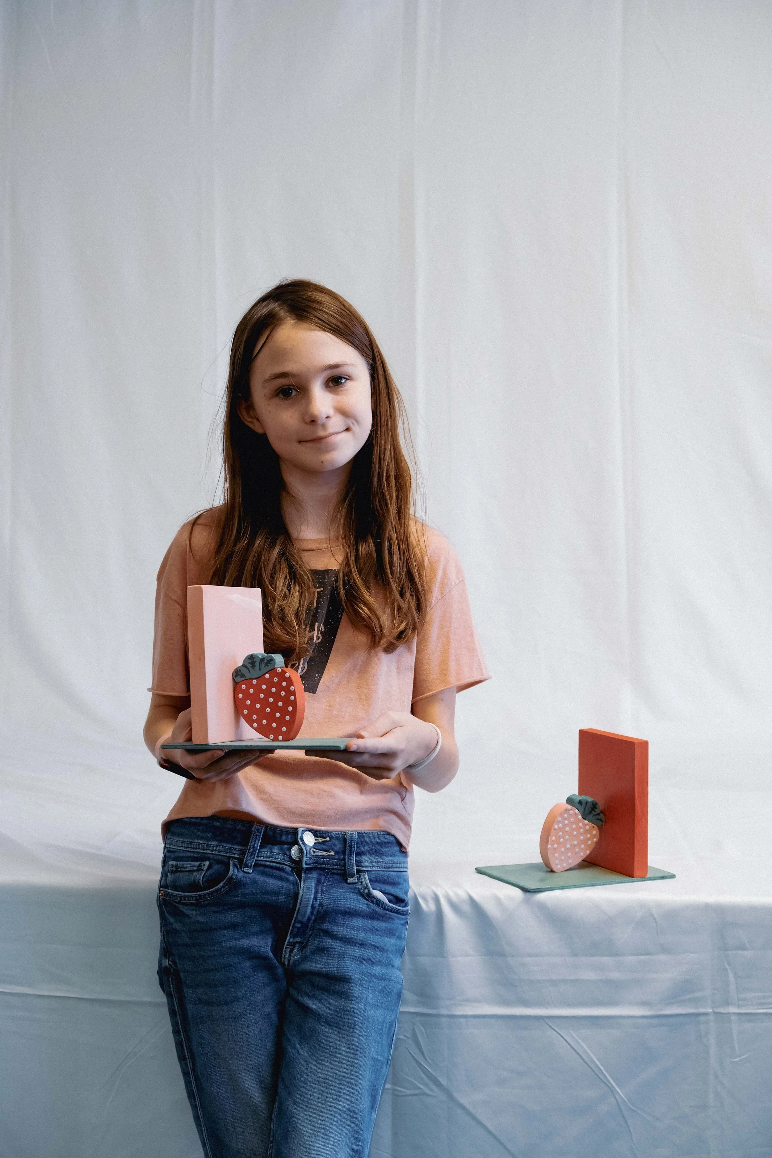 A young girl with long brown hair standing in front of a white background, holding a decorated wooden strawberry craft. There are two more strawberry crafts on a table behind her.