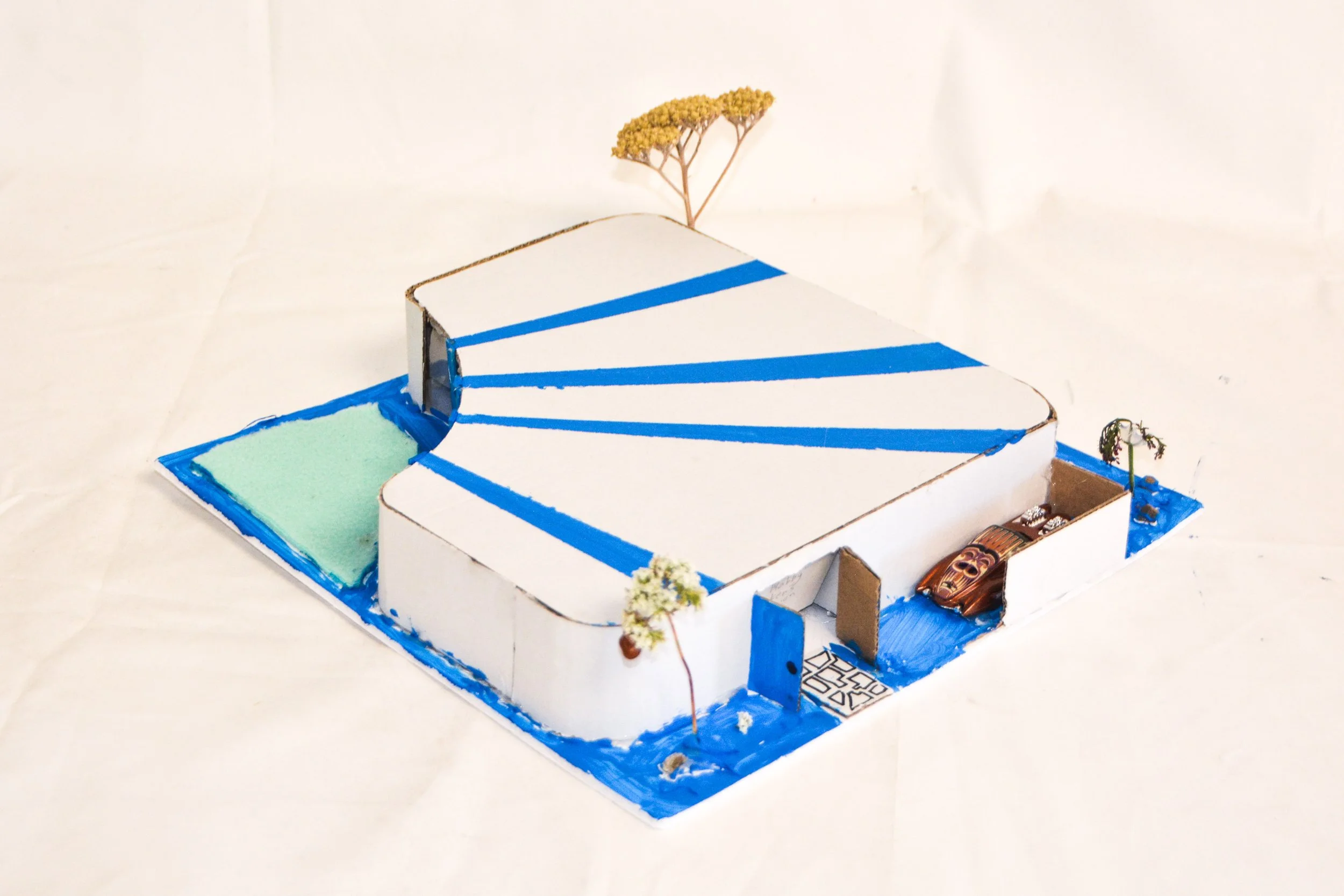 A miniature architectural model of a modern house surrounded by a swimming pool, trees, and pathways, made from foam, cardboard, and modeling materials.