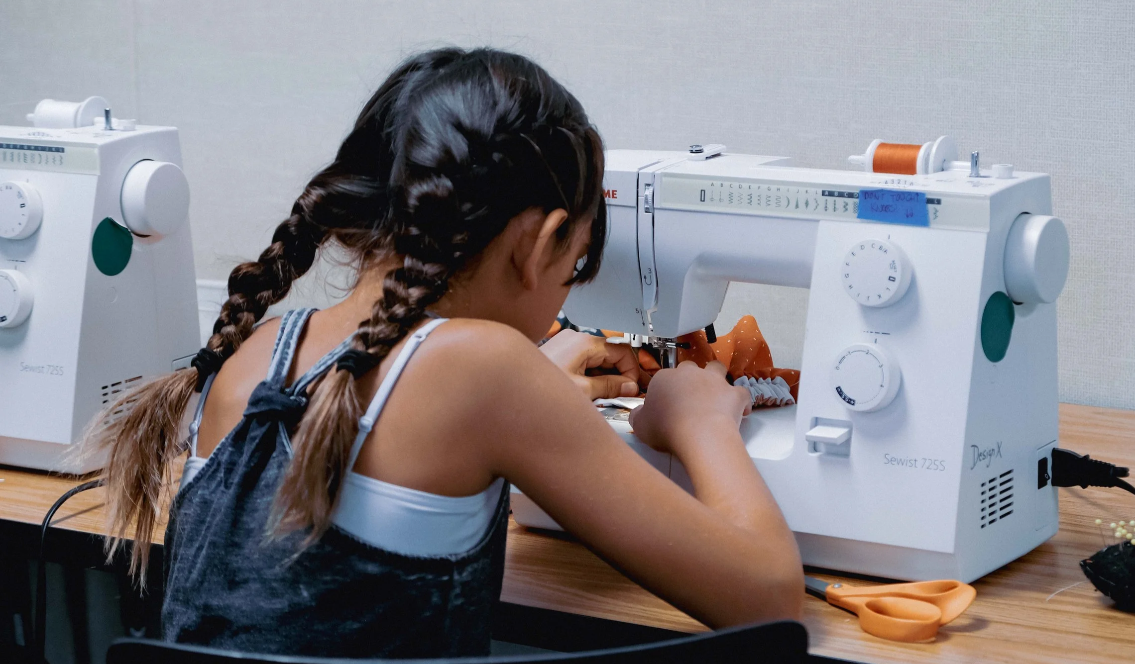 A young girl with braided hair sewing fabric on a white sewing machine at a wooden table.