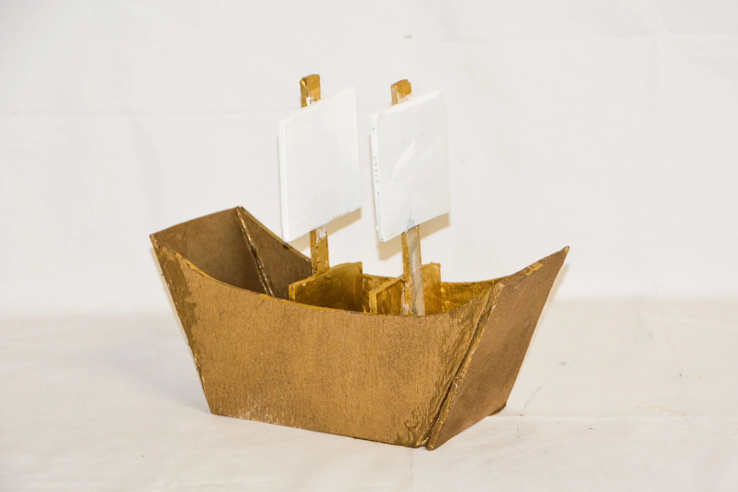 A small handmade boat model with a gold-colored hull and two white sails.