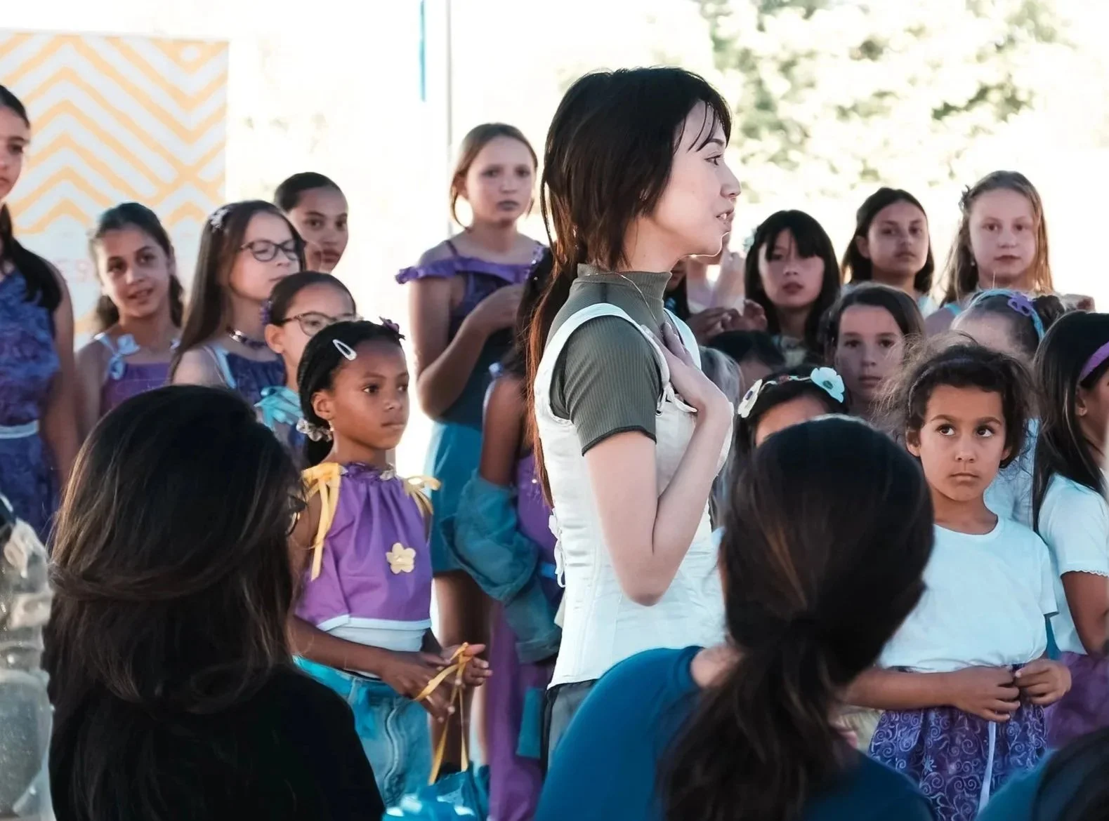 A group of children, mostly girls, are gathered outdoors. A woman is standing in front of them with her hand on her chest, possibly singing or reciting. The children are wearing colorful dresses and look attentive.