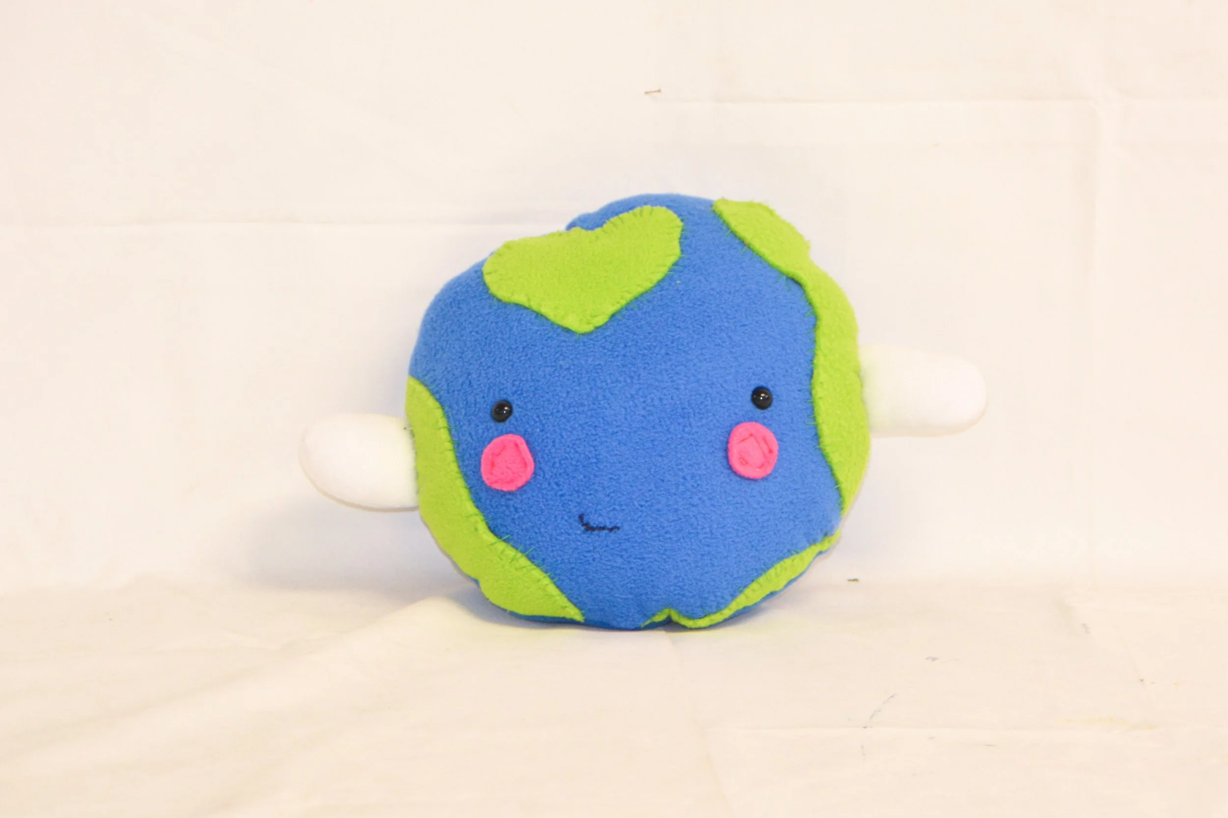 A plush toy shaped like the Earth with a smiling face, pink cheeks, and small black eyes, set against a plain white background.
