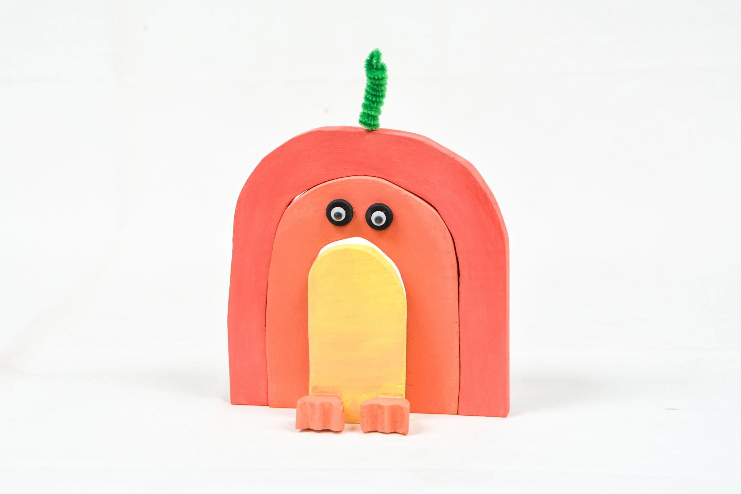 Colorful handmade craft of a penguin inside a pink and orange arch with googly eyes, a yellow belly, and a green pipe cleaner on top, placed on a plain white background.