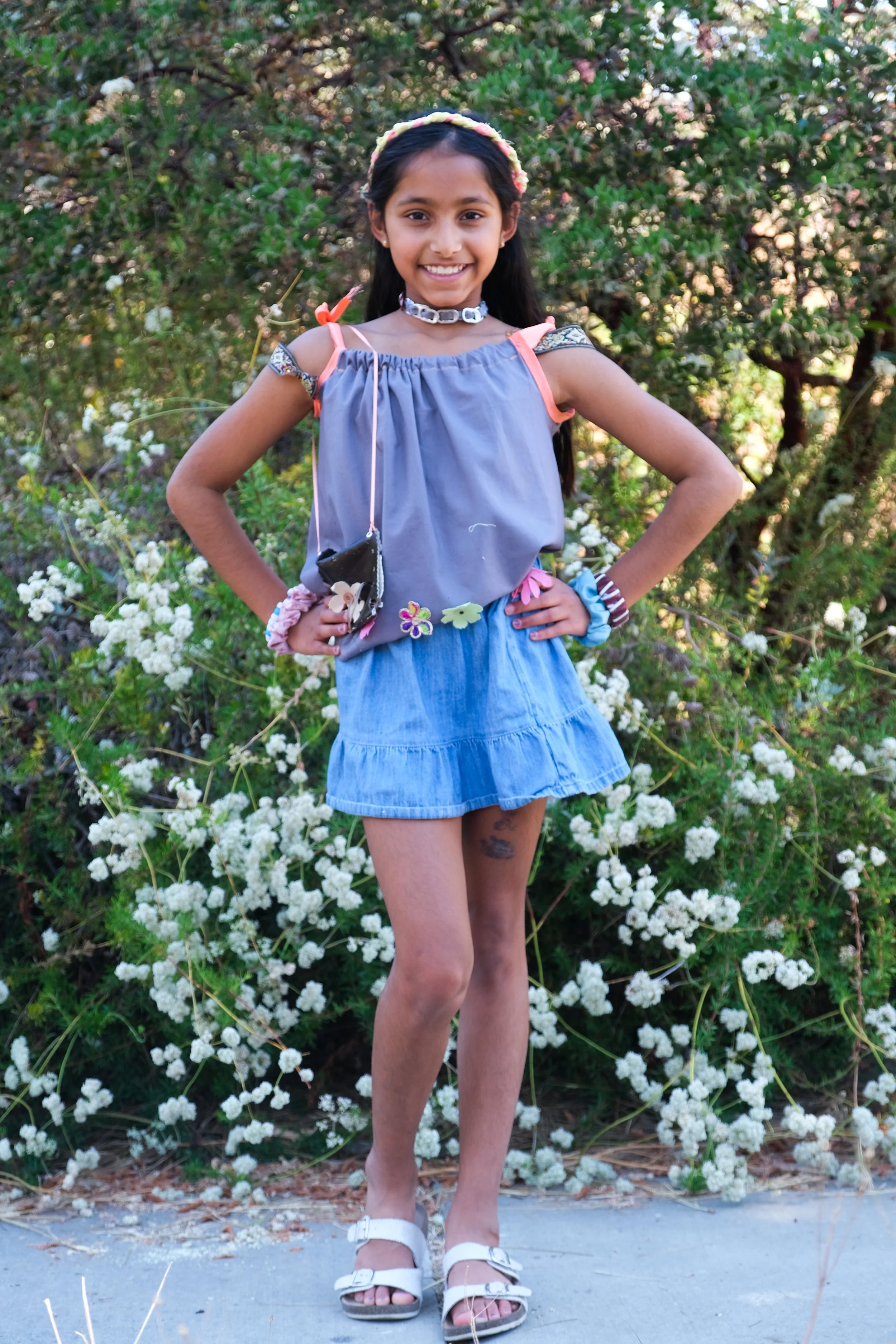 A young girl standing outdoors in front of white flowering bushes, smiling, with hands on her hips, wearing a blue top, denim skirt, white sandals, and colorful accessories.