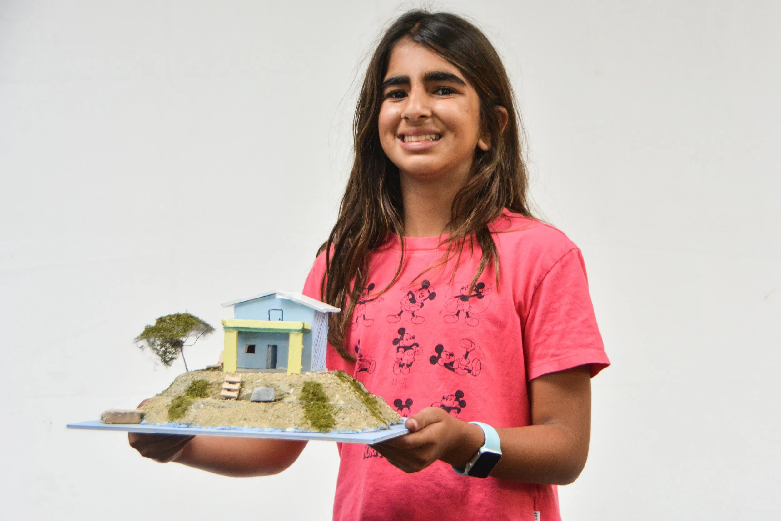 A young girl with long brown hair holding a small model house on a hill, with a tree next to it, smiling at the camera.