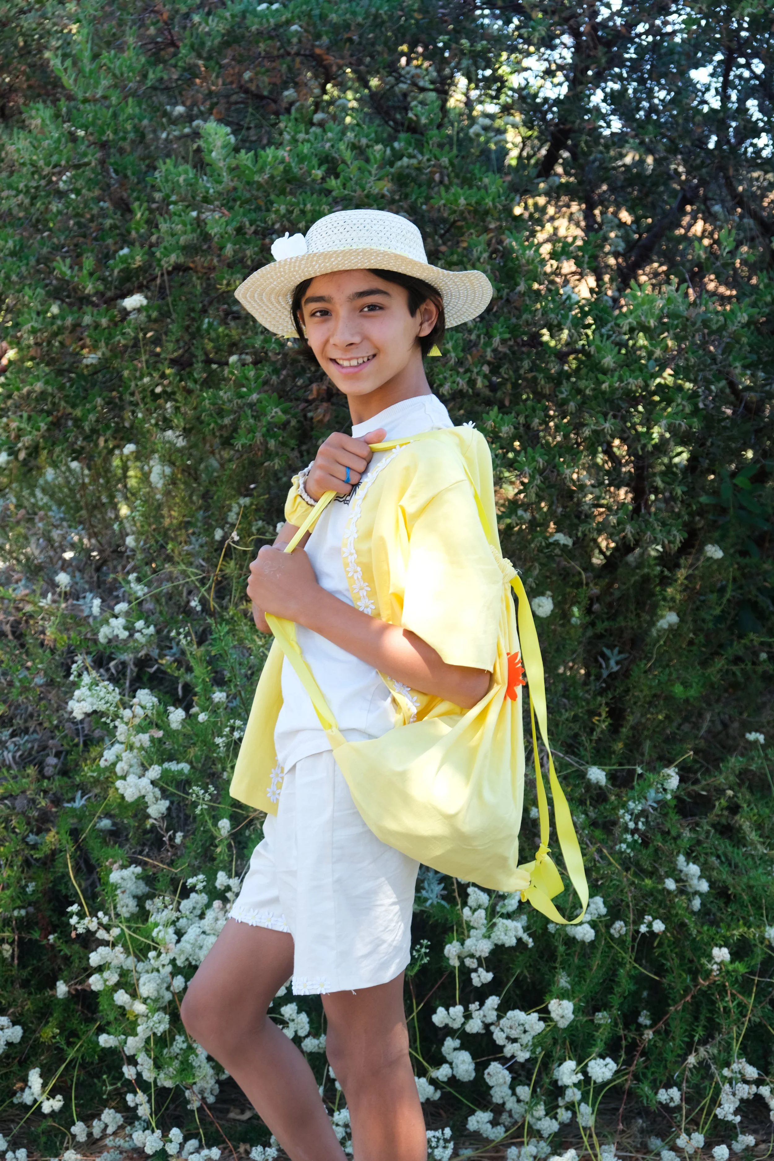 A young girl in a yellow jacket and white shorts, carrying a yellow bag, standing outdoors in front of green bushes with white flowers, wearing a wide-brimmed straw hat and smiling.