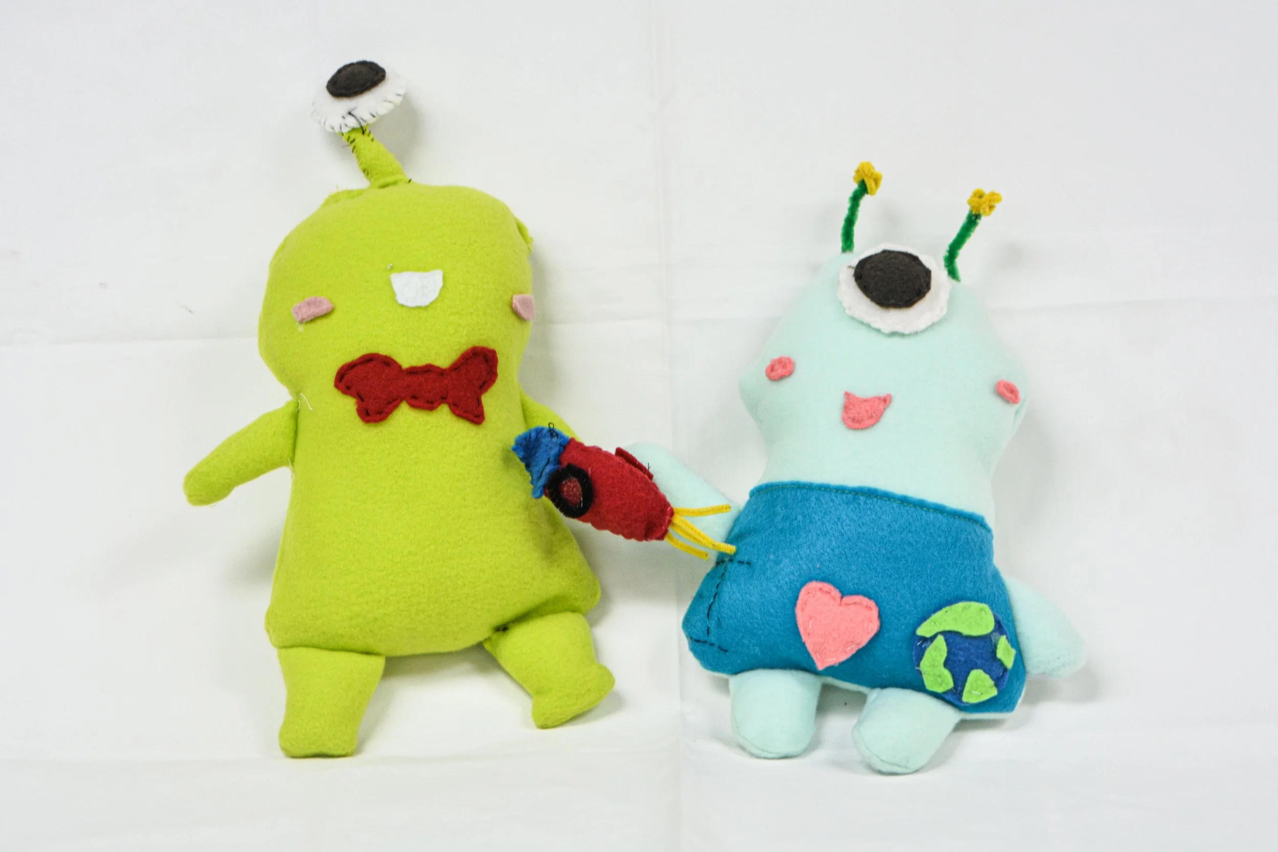 Two handmade plush toys resembling aliens. The green alien has one eye, a red bowtie, and an antenna with a black and white object on top. The light blue alien has two antennas with yellow tips, a large eye, and is holding a small red and blue object