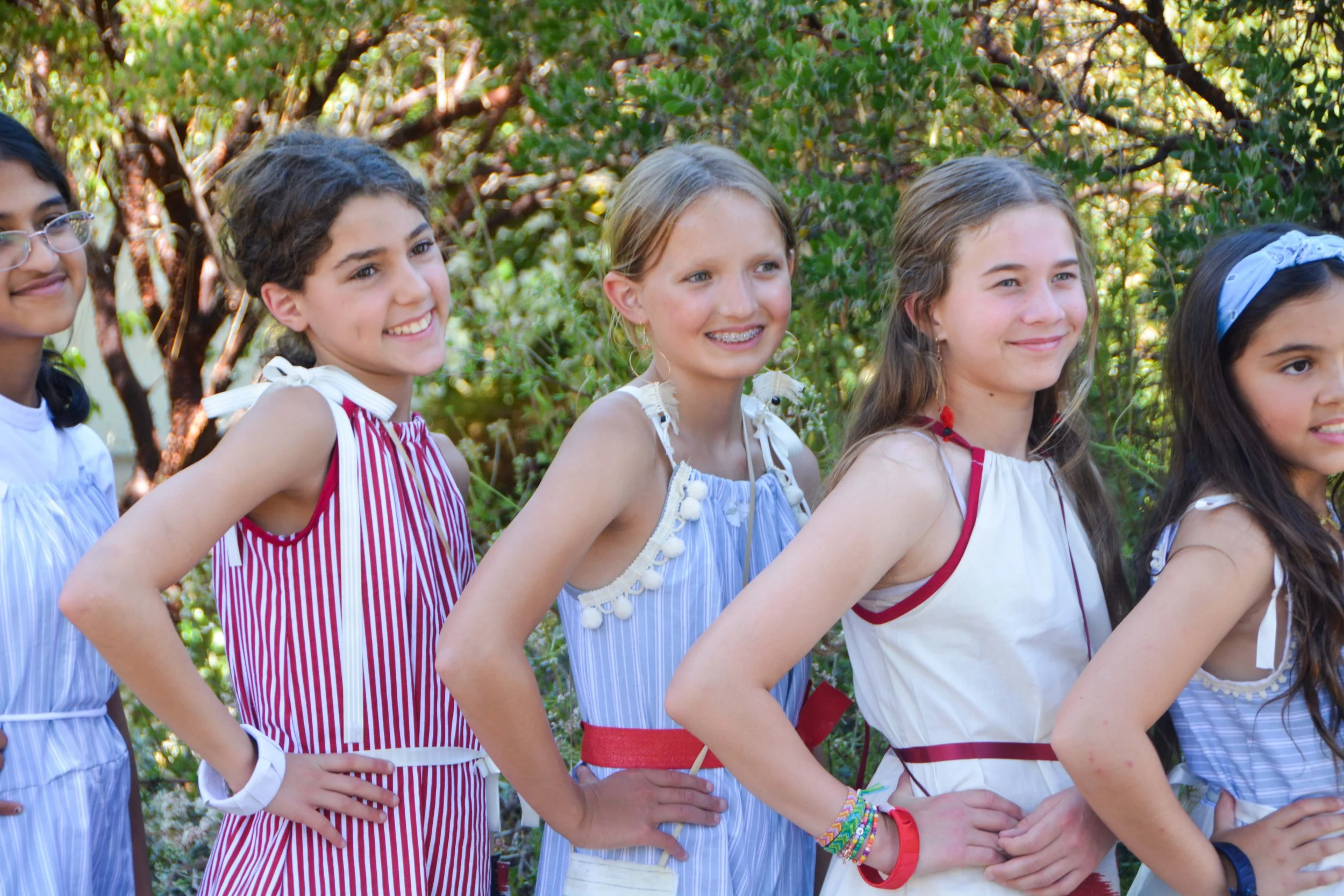 Group of young girls standing outdoors in a line, smiling, with greenery in the background.