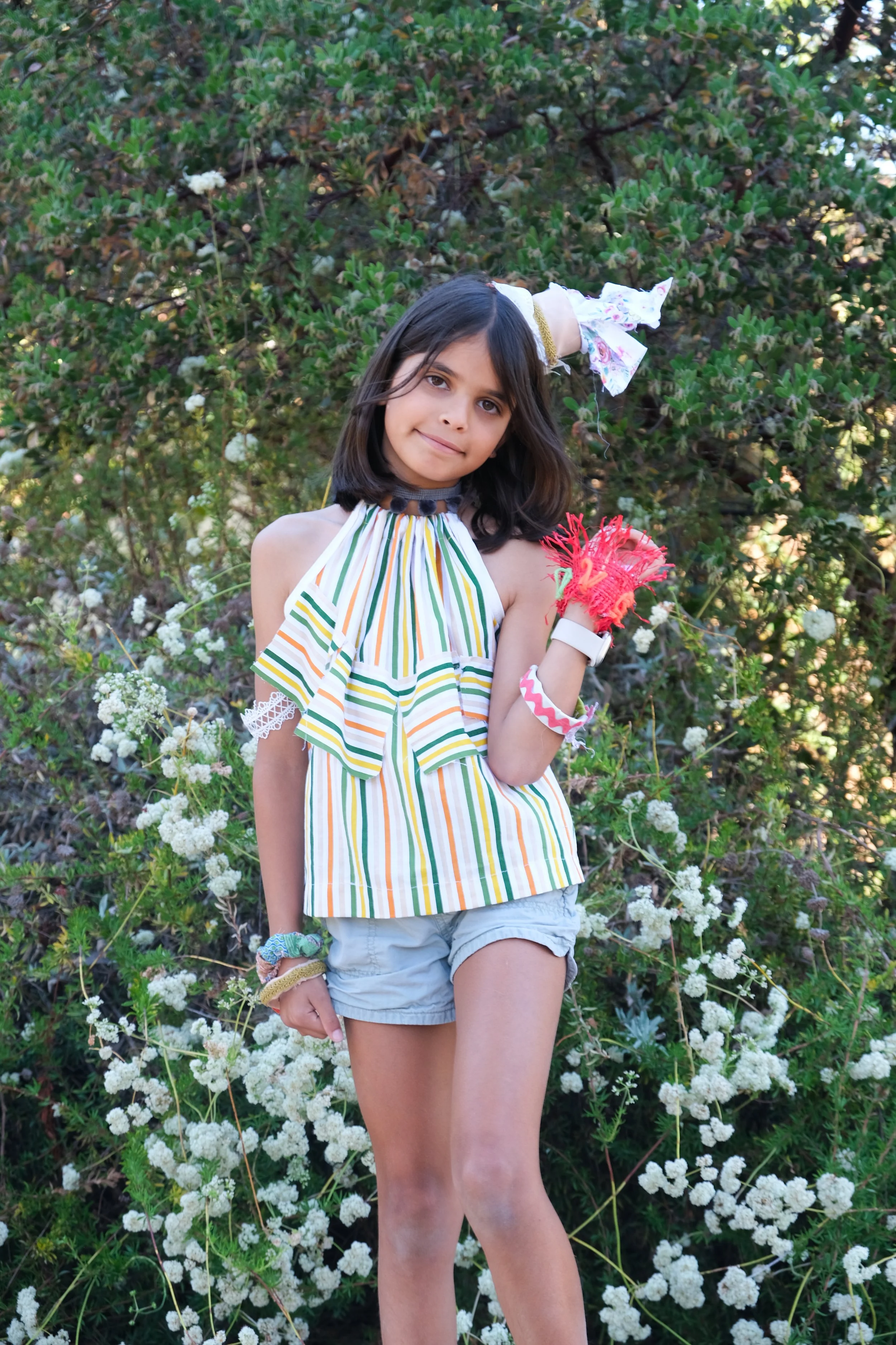 A young girl with shoulder-length dark hair standing outdoors in front of green bushes with white flowers, wearing a colorful striped top, light-colored shorts, and various accessories, including a bow, gloves, and bracelets.