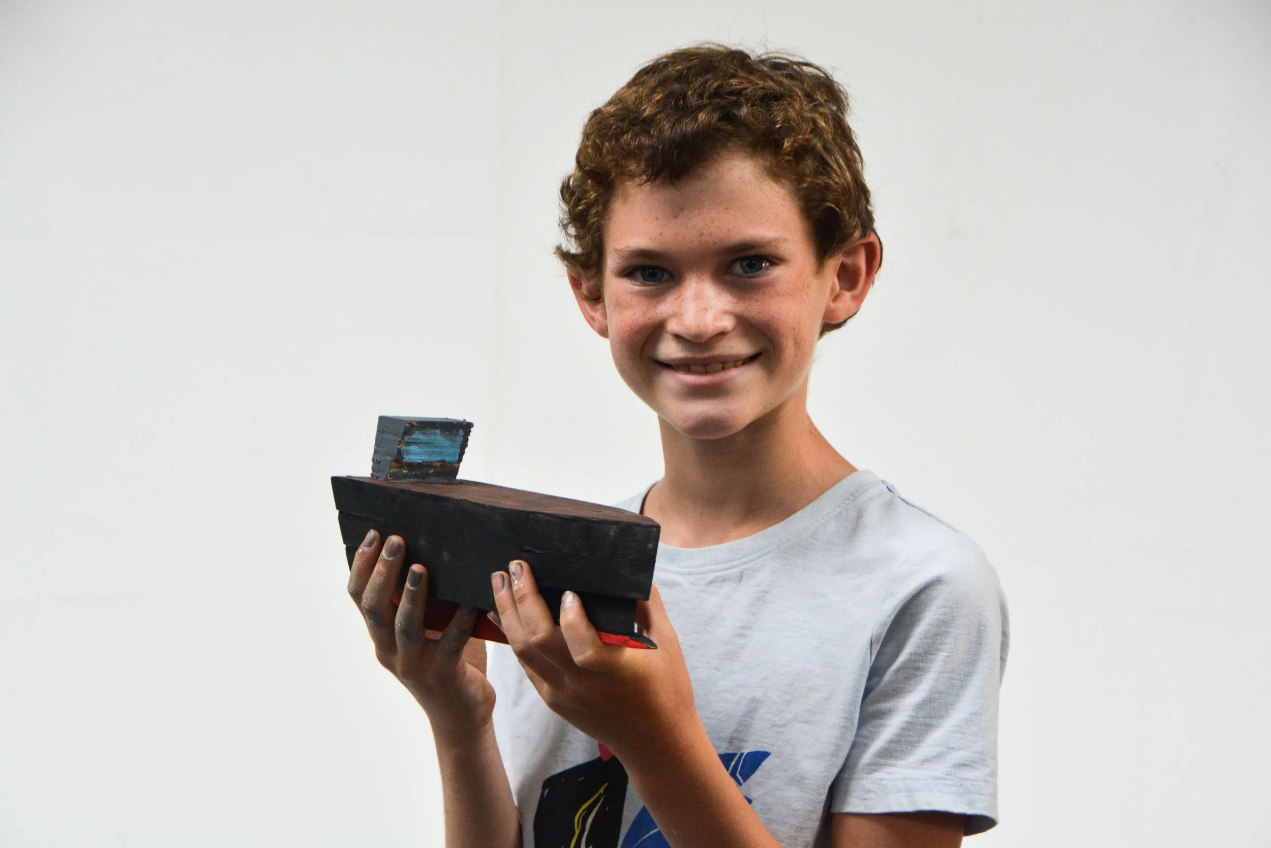 A young boy with curly brown hair and freckles smiling while holding a small wooden boat model painted in dark colors.