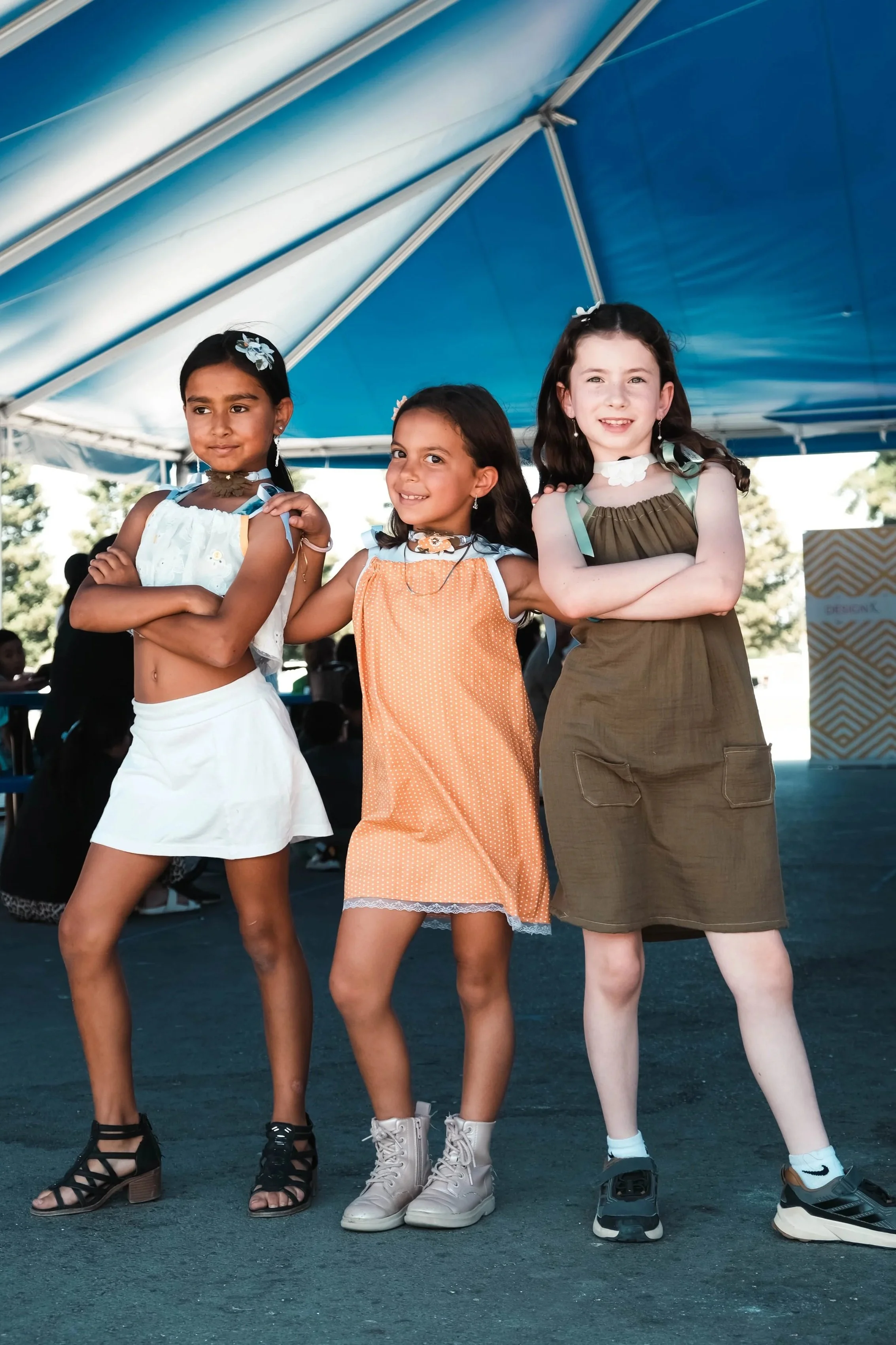 Three young girls with arms crossed, standing under a large blue tent at an outdoor event.