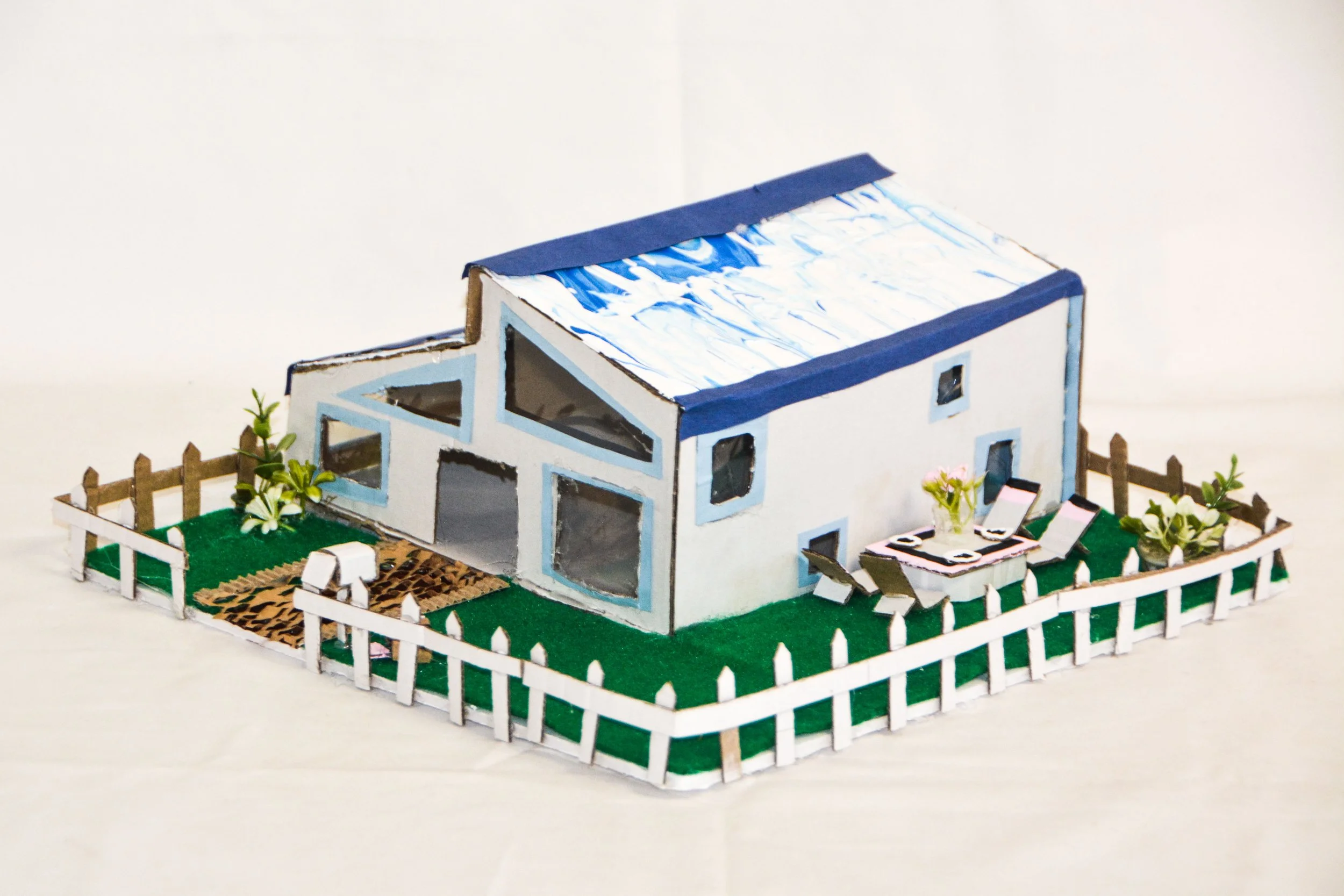 A miniature model of a modern house with a backyard, white picket fence, outdoor table with chairs, and small garden areas.