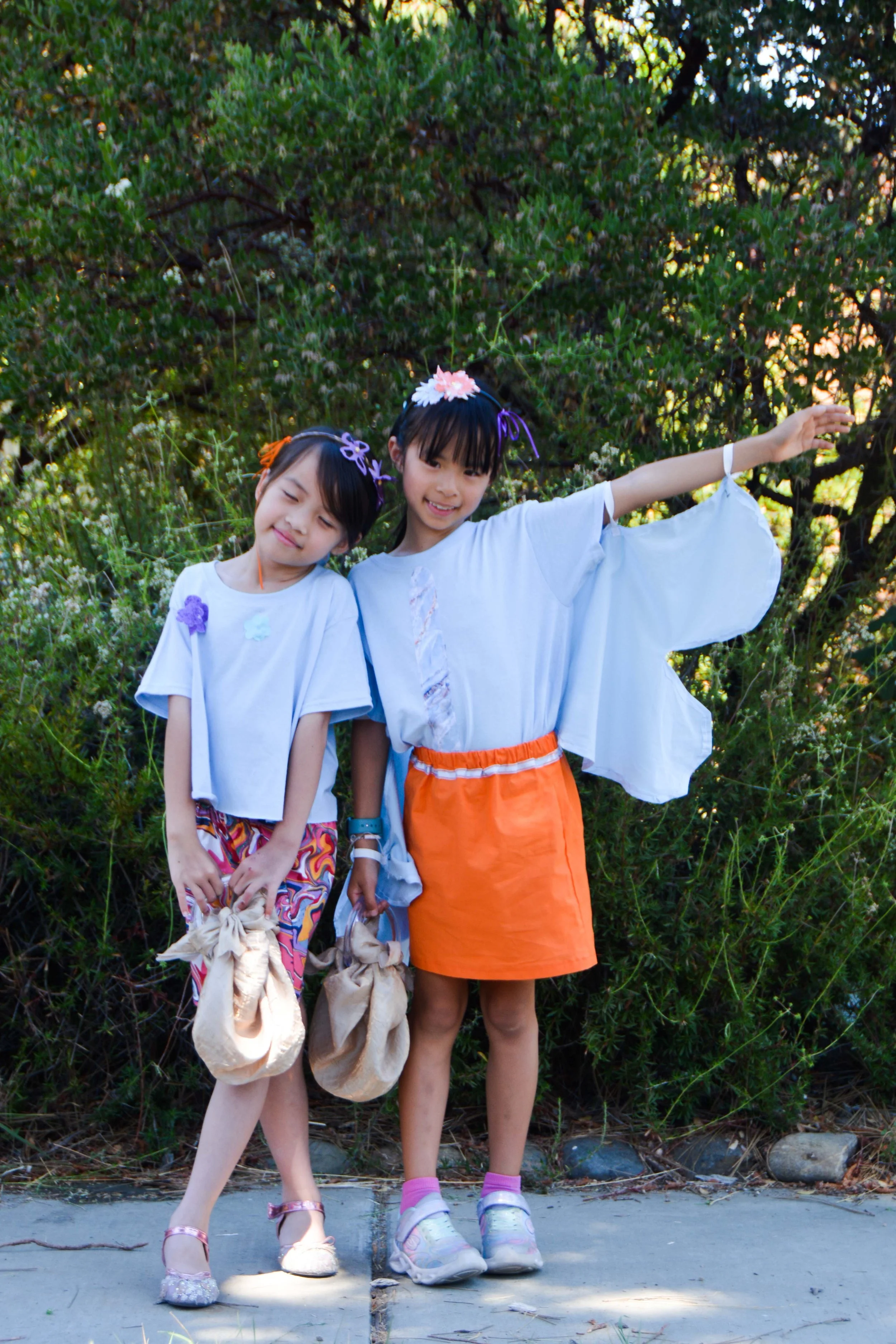 Two young girls in colorful summer outfits standing outdoors in front of green foliage, holding beige bags, with the girl on the right extending her arm outward and both smiling.