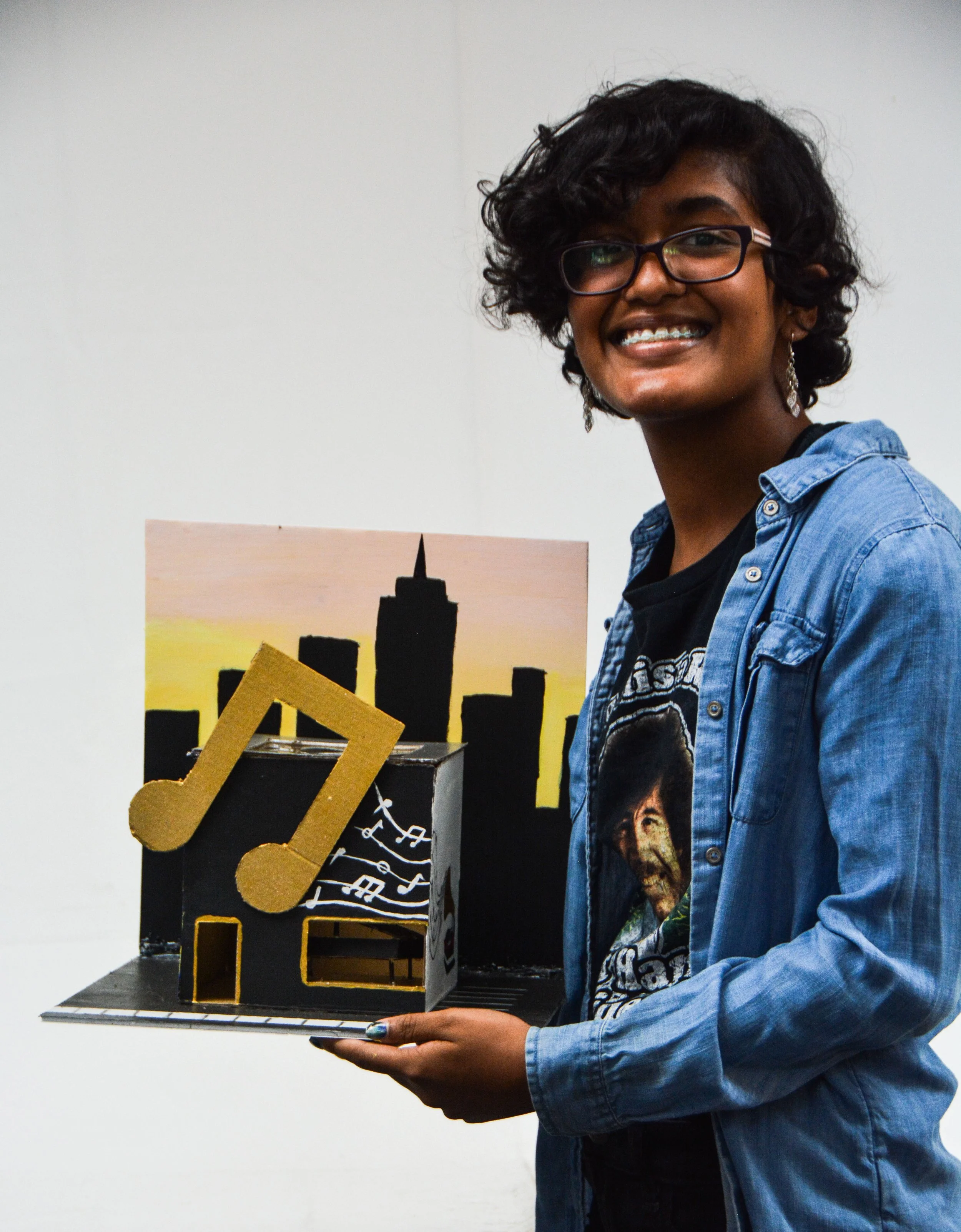 A person smiling and holding a small, decorated model of a music-themed building with a city skyline in the background.