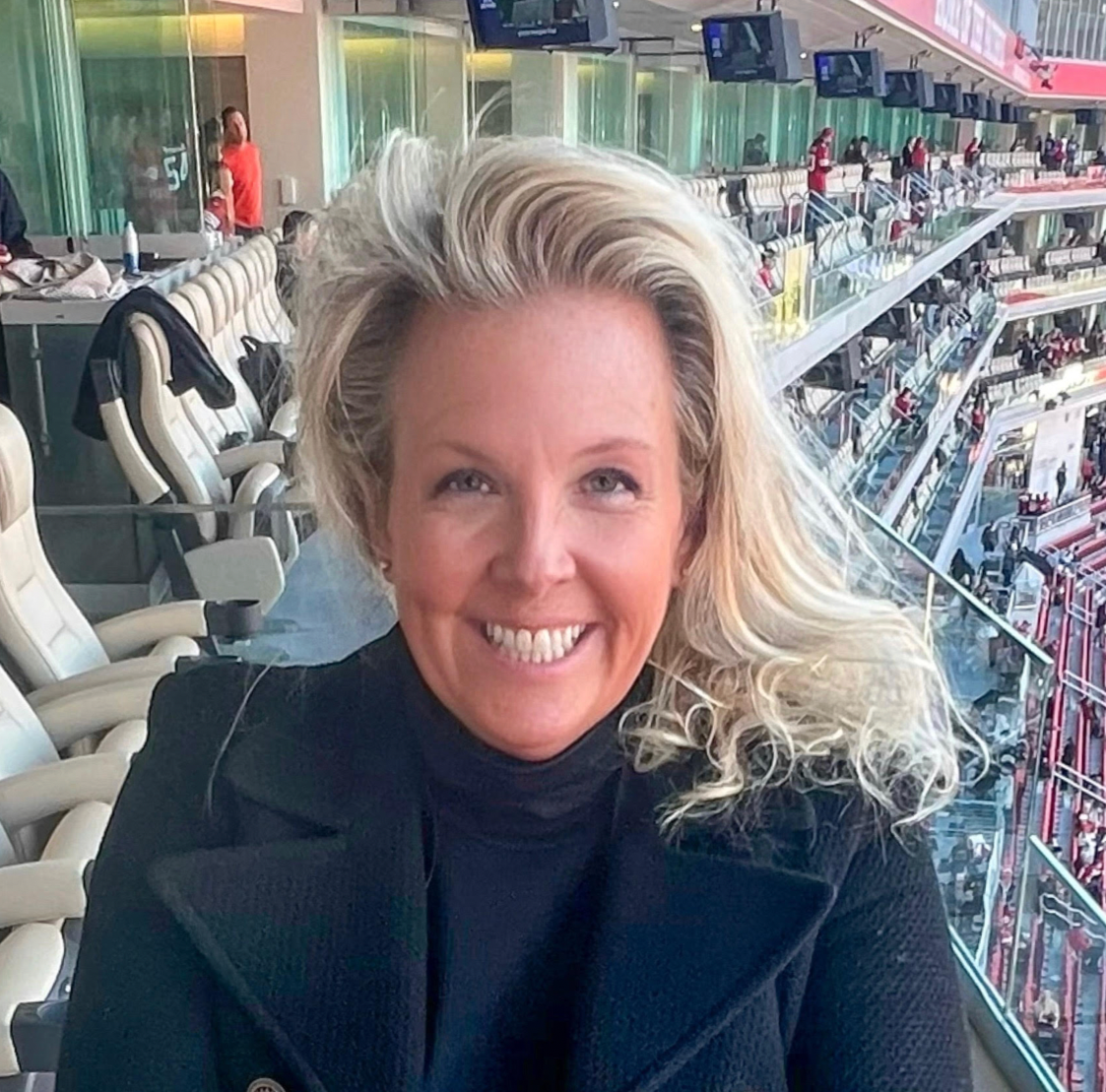 A woman with blonde curly hair smiling at the camera in a stadium or arena with multiple levels and other people in the background.