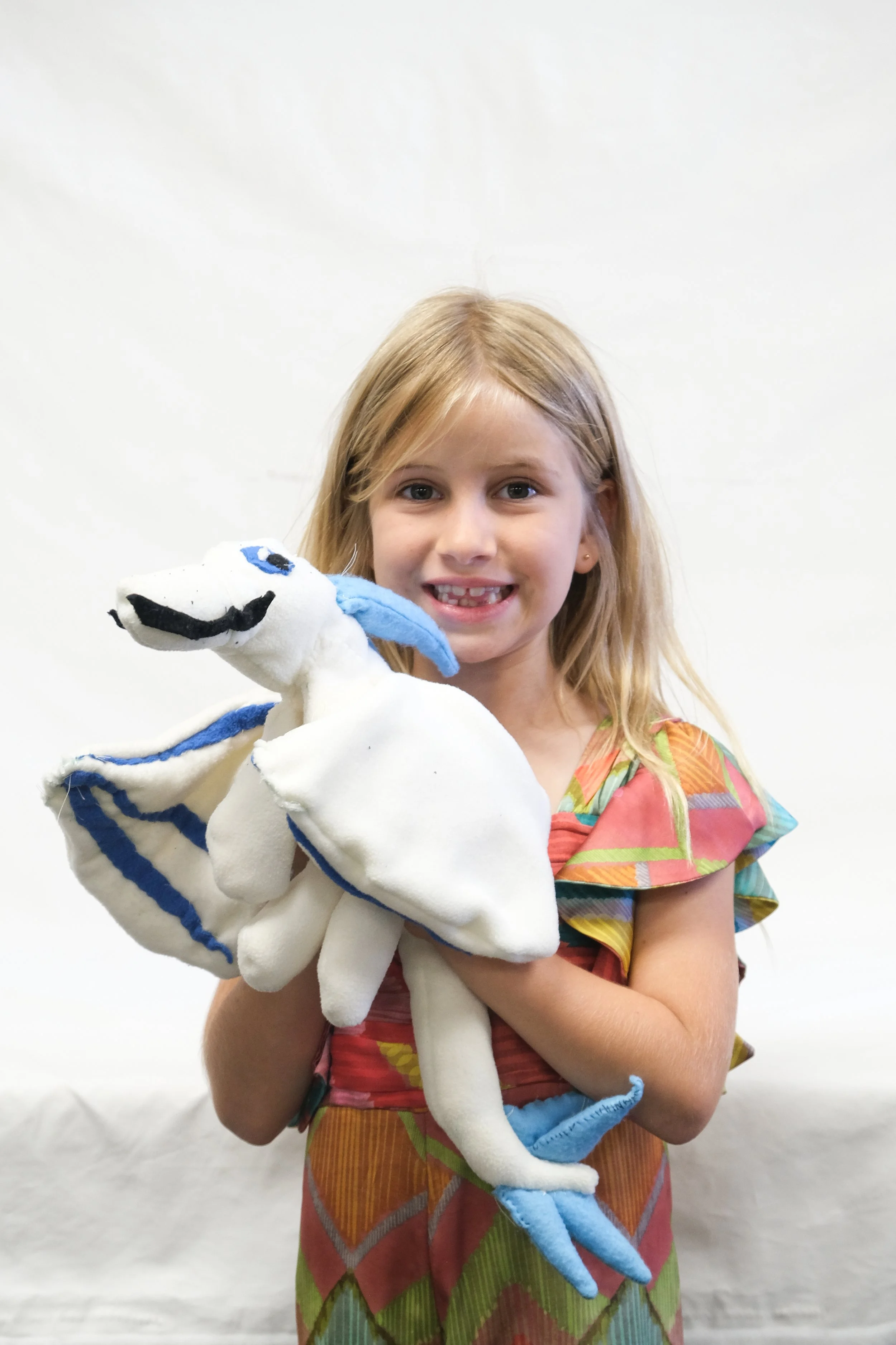 A young girl with blonde hair holding a white plush toy dolphin, smiling at the camera against a plain white background.