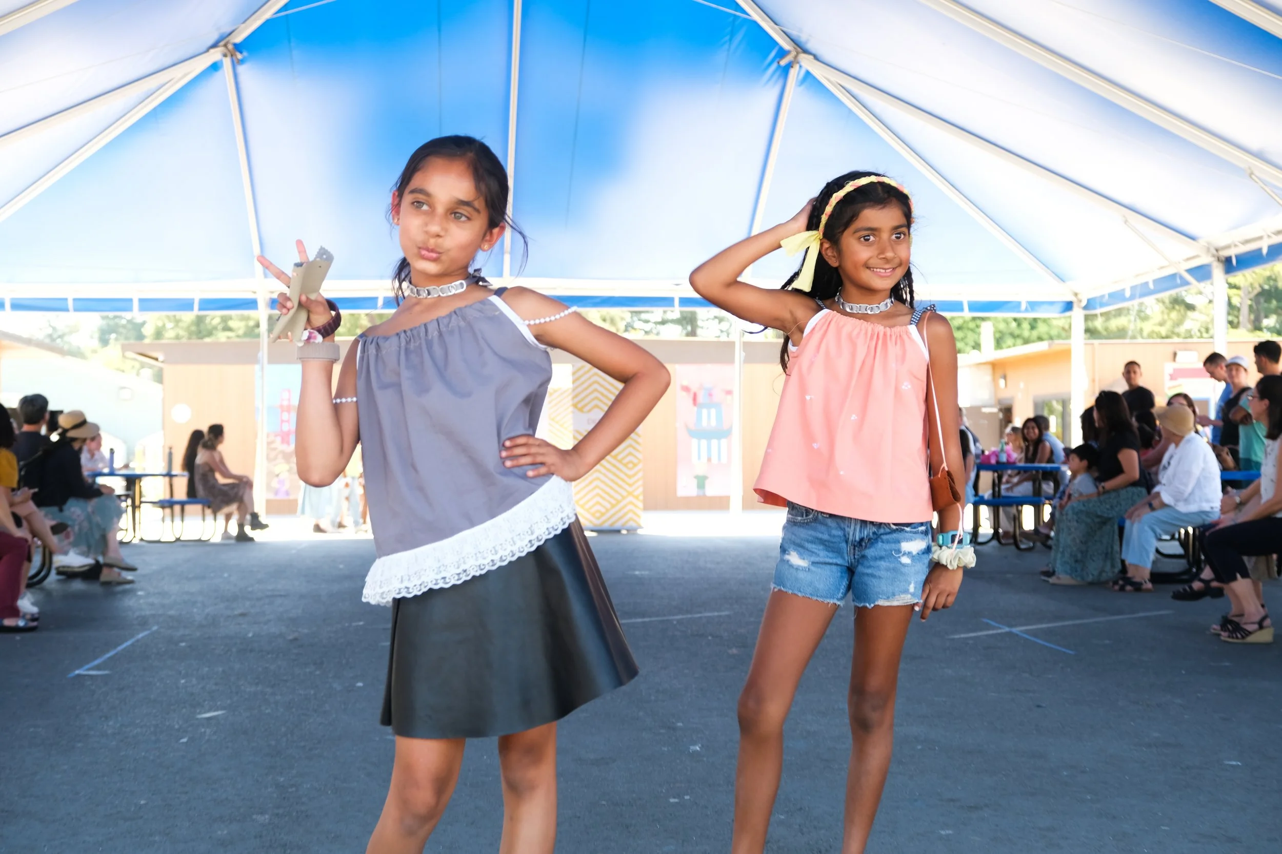 Two young girls posing under a large tent at an outdoor event, with seated spectators in the background.
