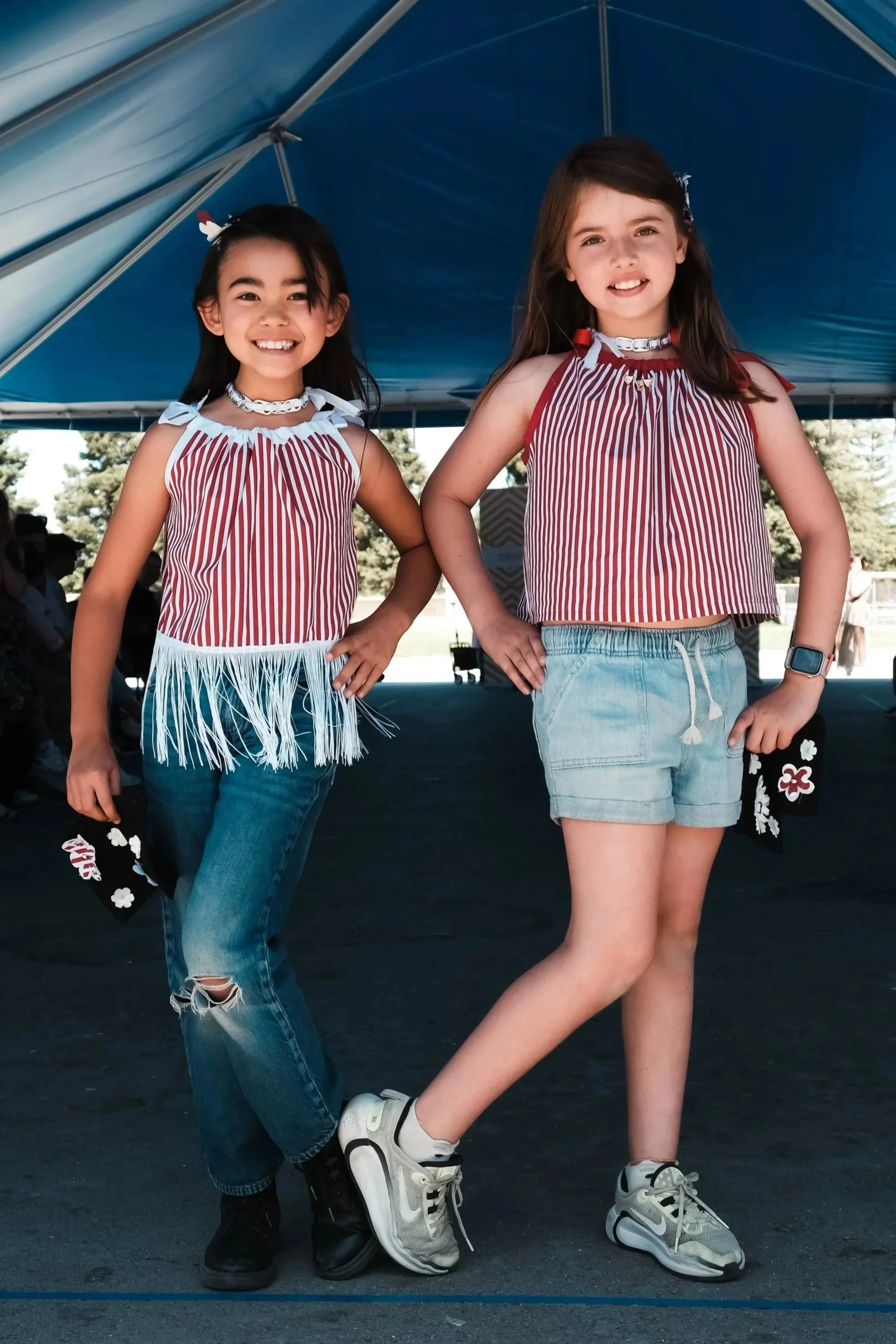 Two young girls standing under a blue canopy, smiling and posing for the photo, dressed in casual summer clothes.