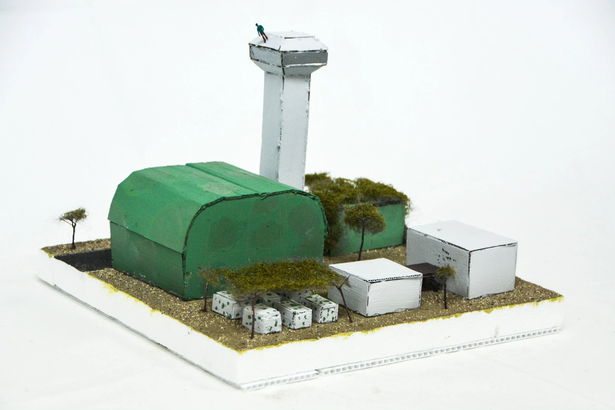 Miniature model of an industrial farm with a large green greenhouse, two white storage buildings, trees, and a tall control tower.
