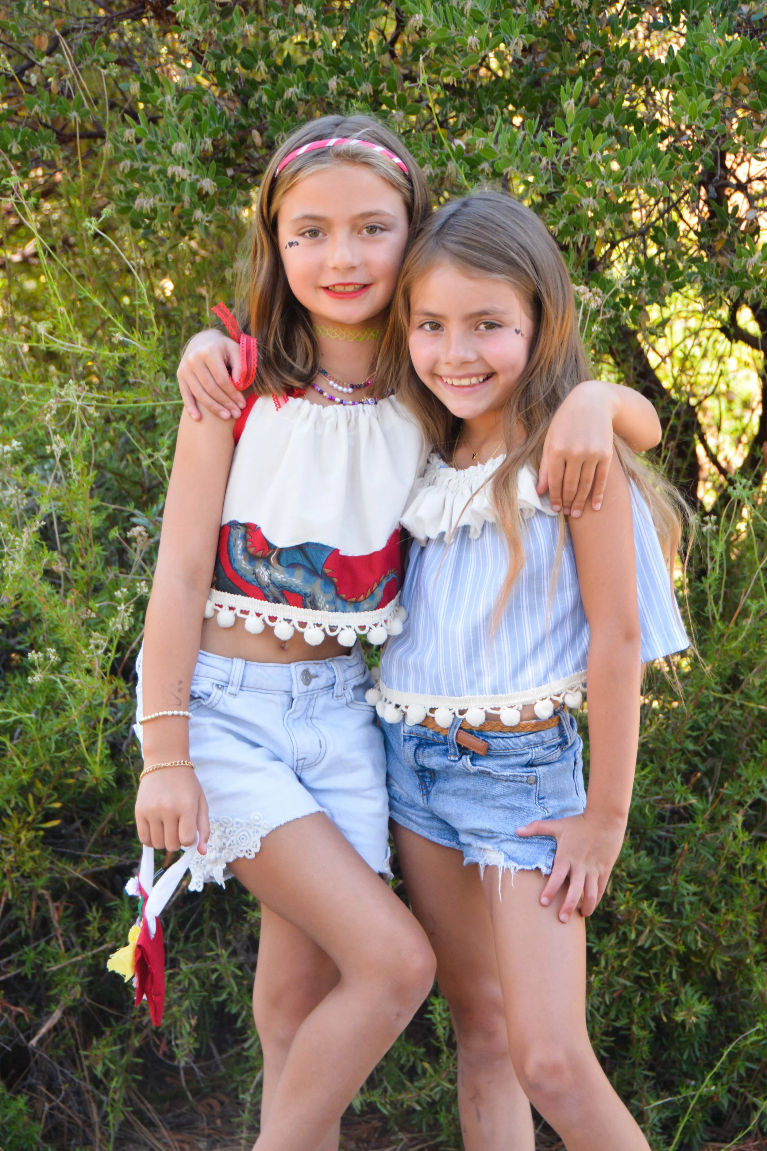 Two young girls standing outside in front of green bushes, smiling and embracing each other.