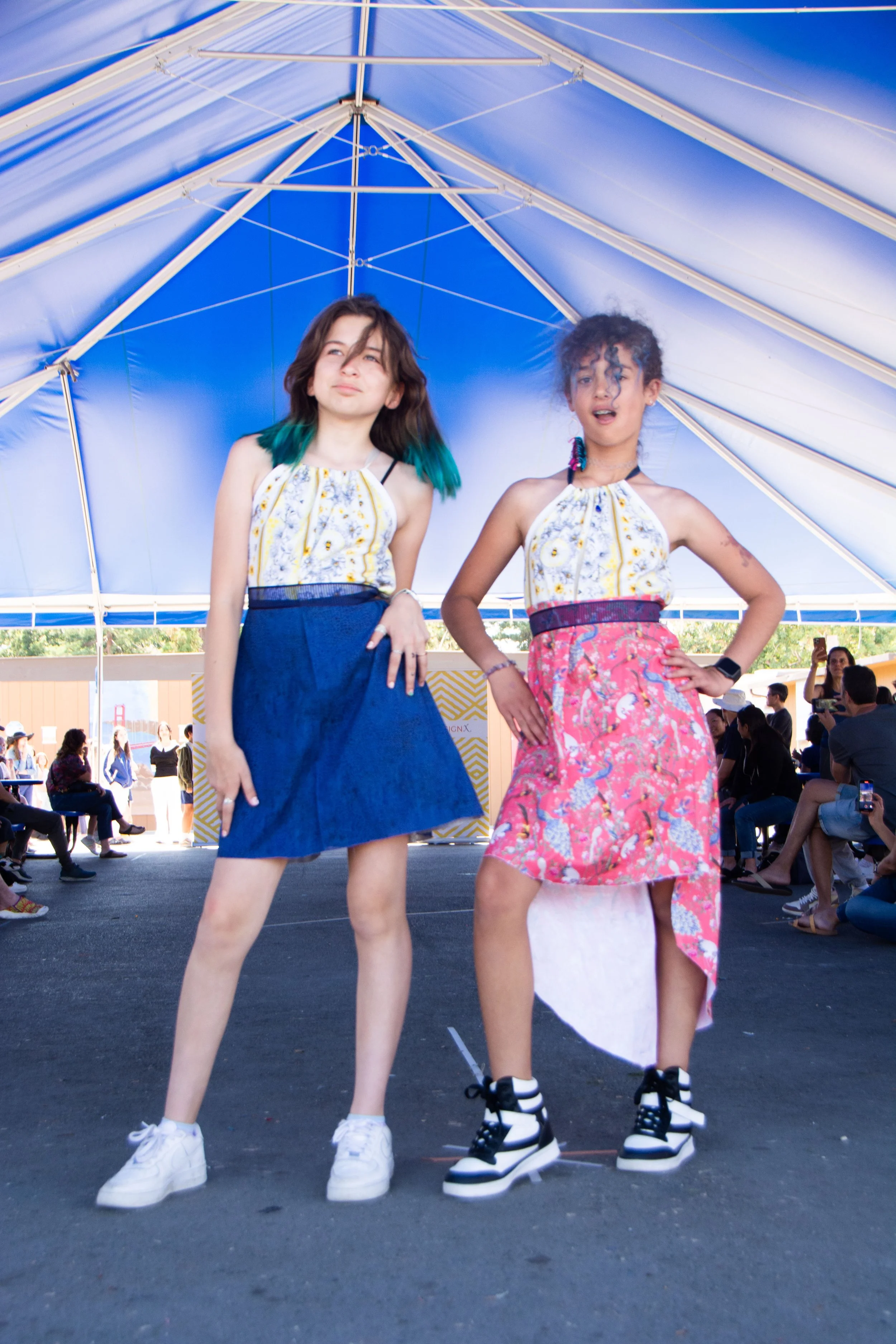 Two young women posing in colorful outfits under a blue and white canopy at an outdoor event, with seated and standing people in the background.