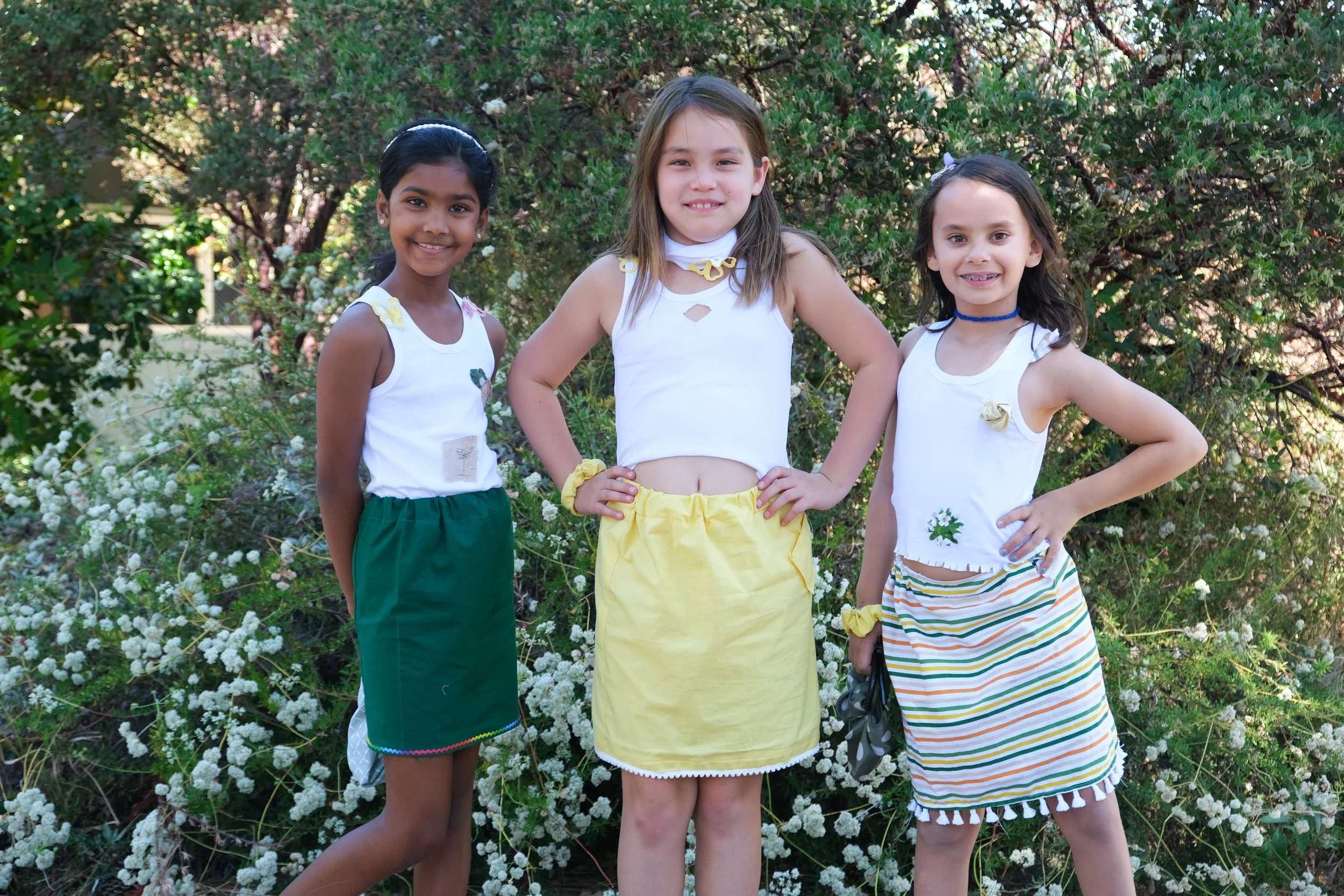 Three young girls standing outdoors in front of green bushes with white flowers, dressed in summer outfits and smiling at the camera.