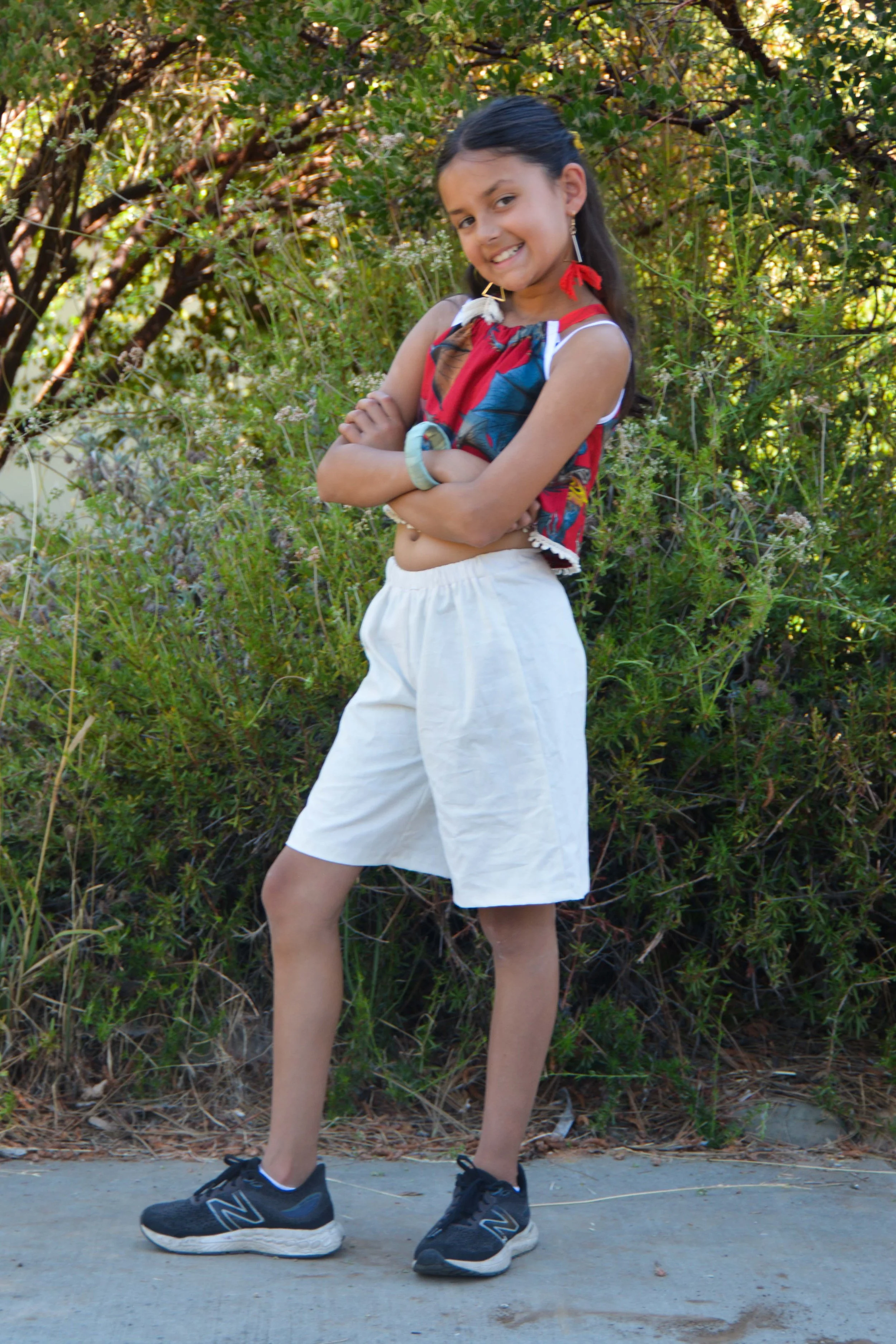 A girl smiling with arms crossed, wearing a colorful sleeveless top, white shorts, black sneakers, earrings, and a bracelet, standing outdoors in front of green bushes.