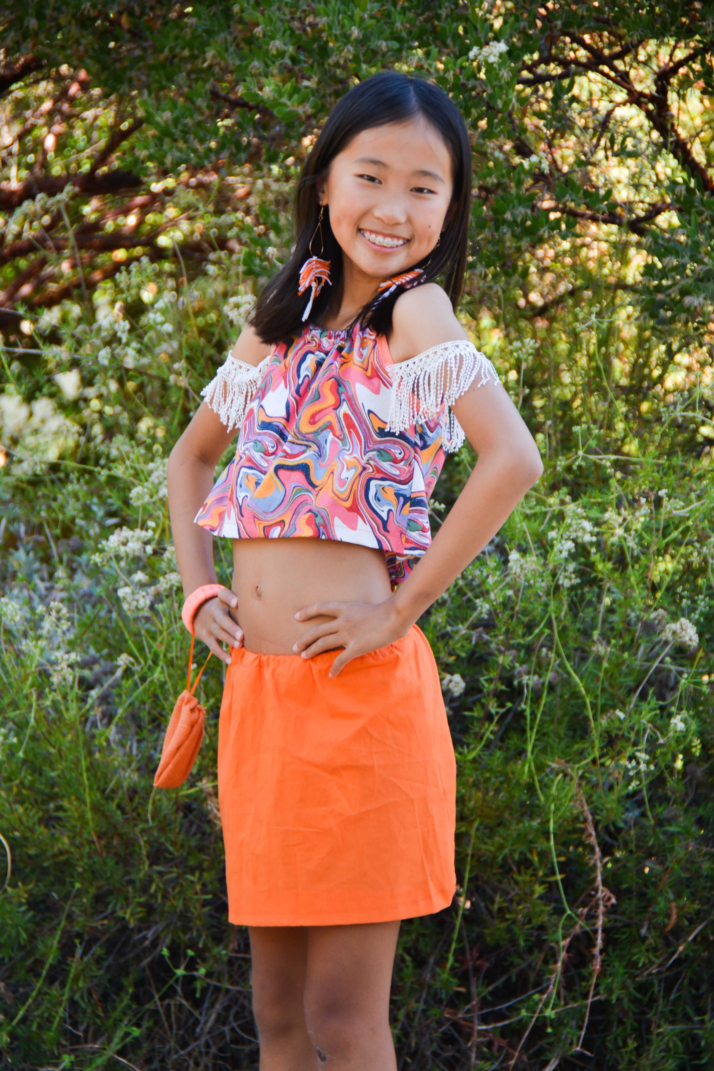 A girl with long black hair smiling outdoors, wearing an off-shoulder top with fringe details, bright orange skirt, and vibrant earrings, standing in front of lush green foliage.