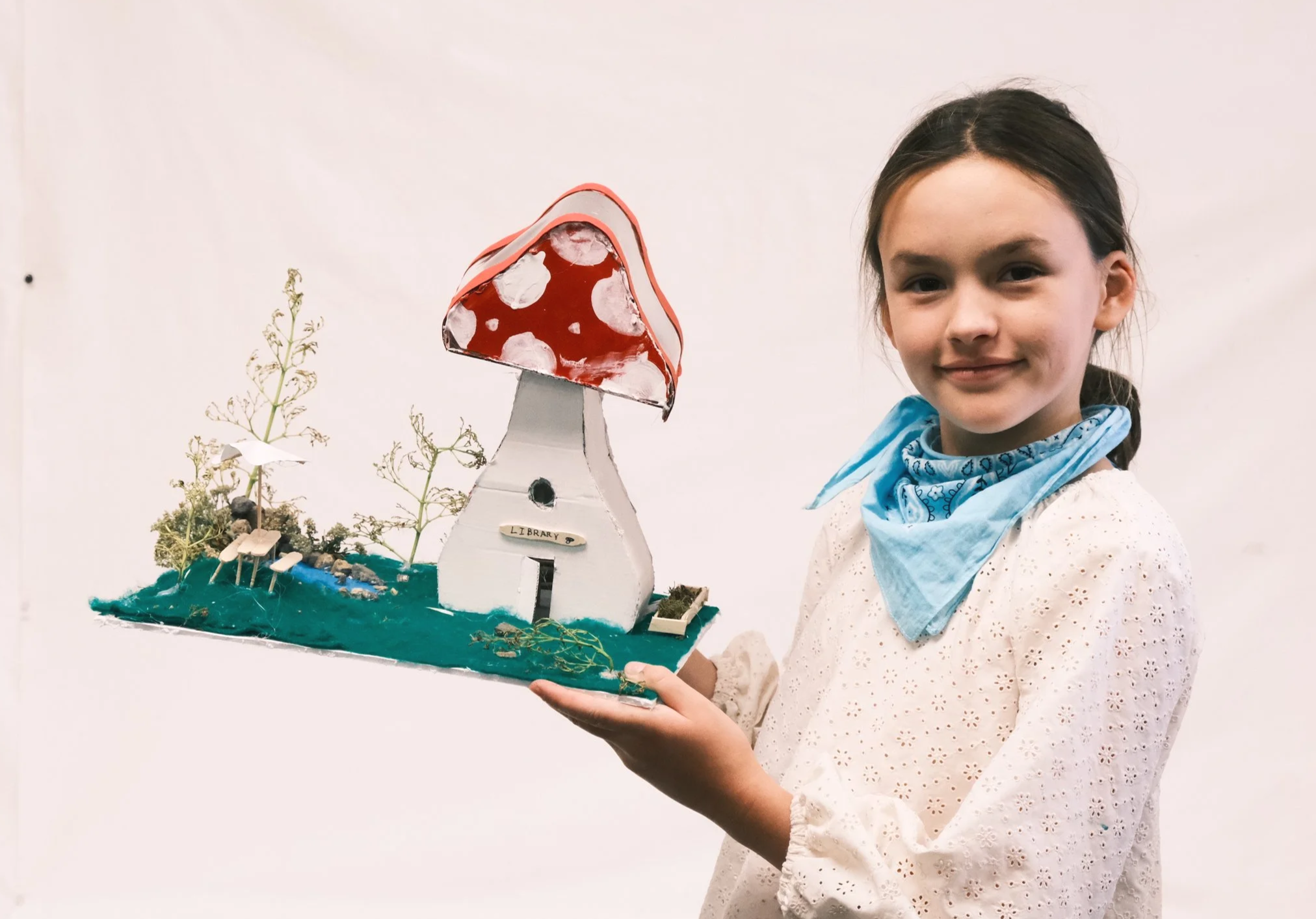 A young DesignX student with dark hair and a blue bandana around her neck, holding a model of a mushroom-shaped library building, with a large red mushroom cap roof, a small door, and surrounding greenery.