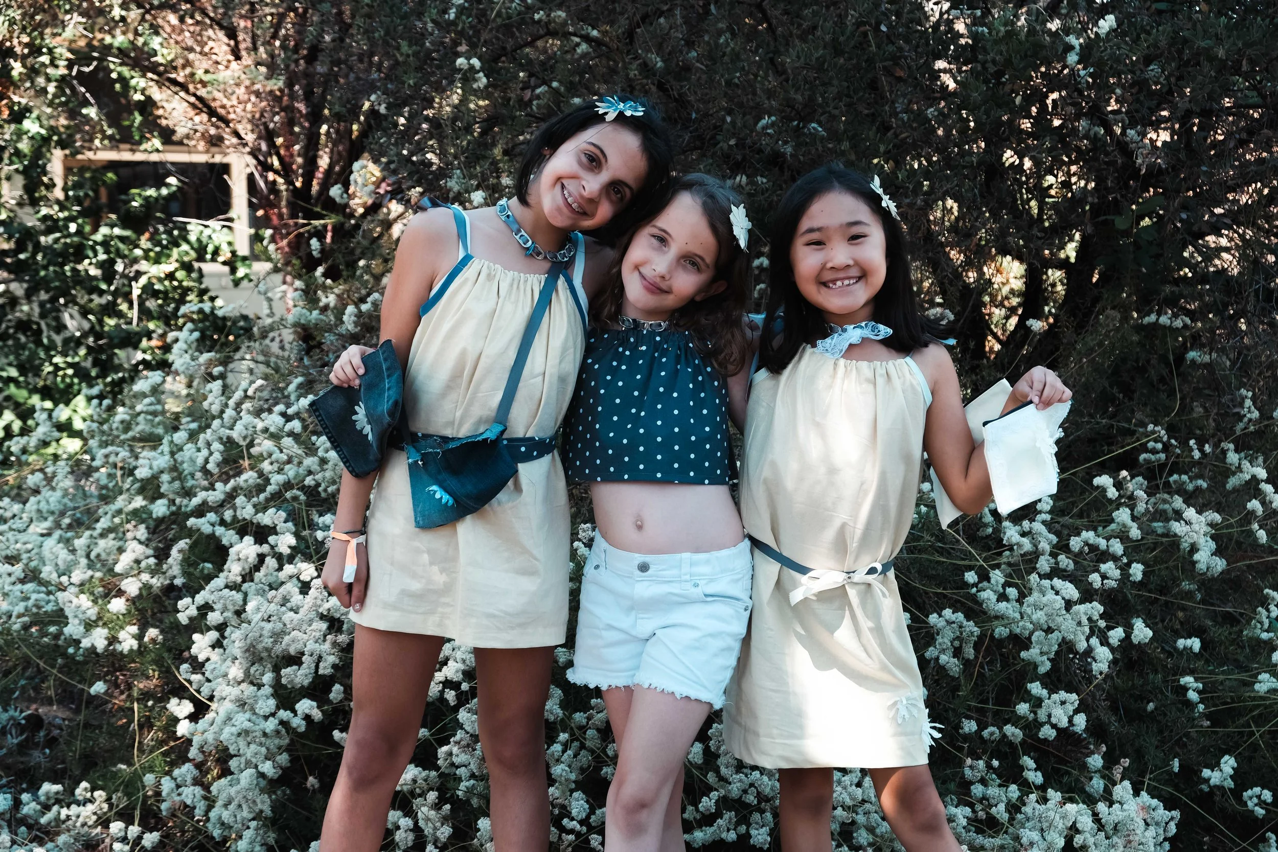 Three girls standing in front of a bush with white flowers, smiling and posing for the camera.