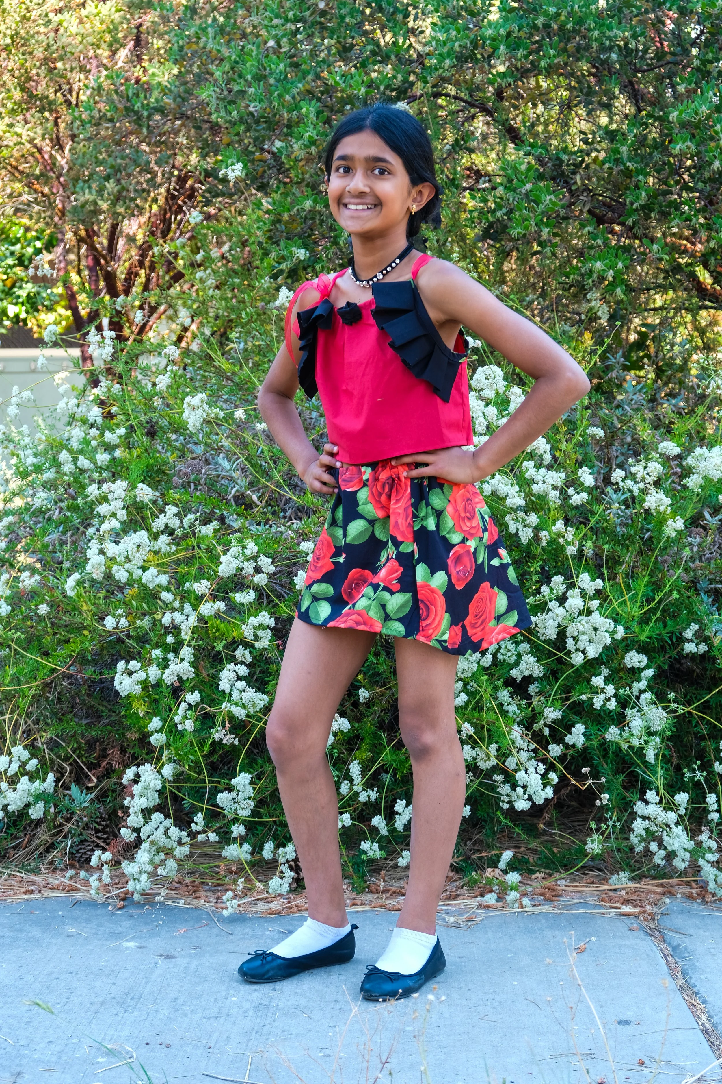 A young girl smiling and posing outdoors in front of green bushes with white flowers, wearing a red top with black bows, a floral skirt with red roses and green leaves, white socks, and black ballet flats.